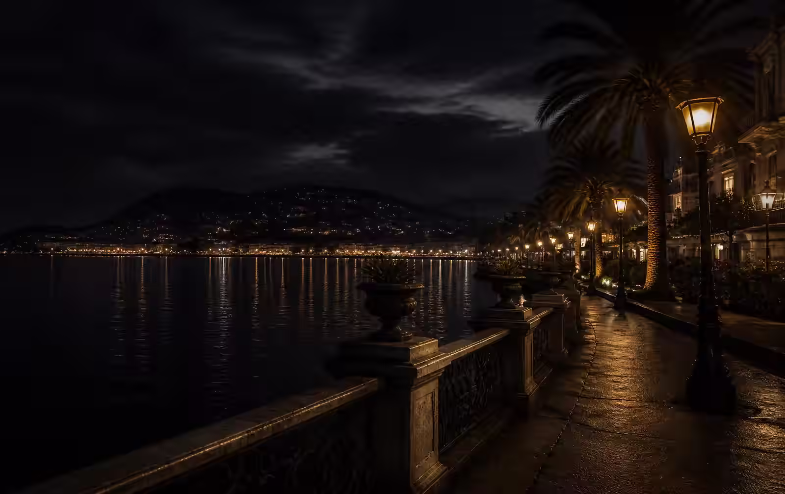 A waterfront city promenade at night beside a dark bay with palms and warm lamplight