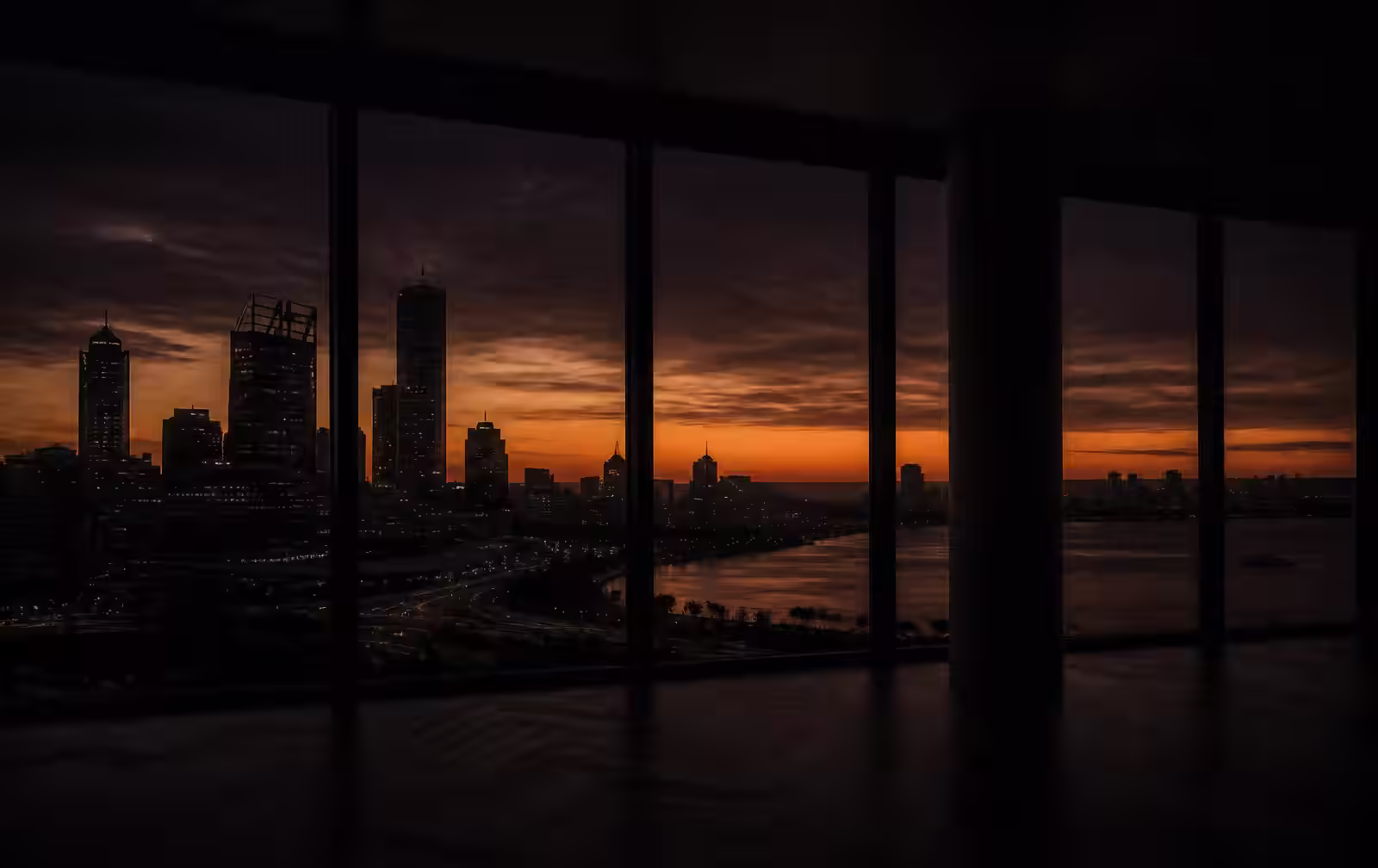 Perth's skyline and river at deep dusk with a burnt-orange horizon seen from an unlit modern interior