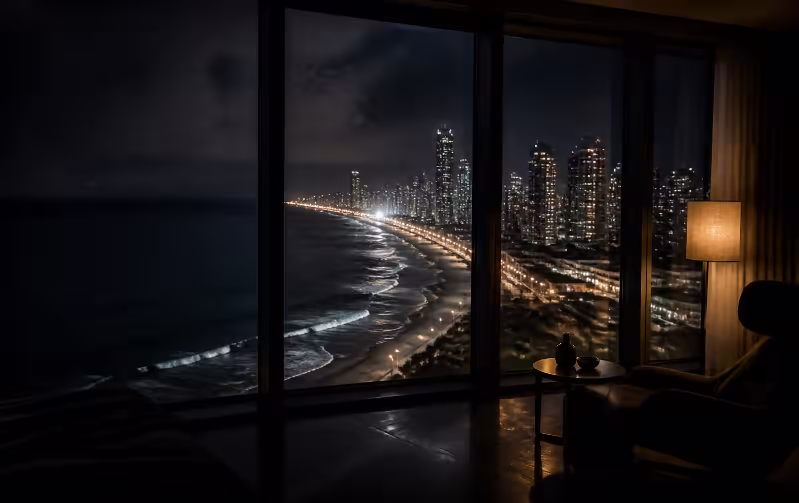 A lit coastal high-rise skyline and dark surf seen at night from an unlit modern apartment interior