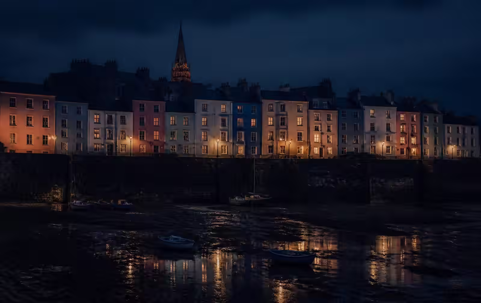 Pastel Welsh harbour houses lit at night above a dark tidal harbour with moored boats
