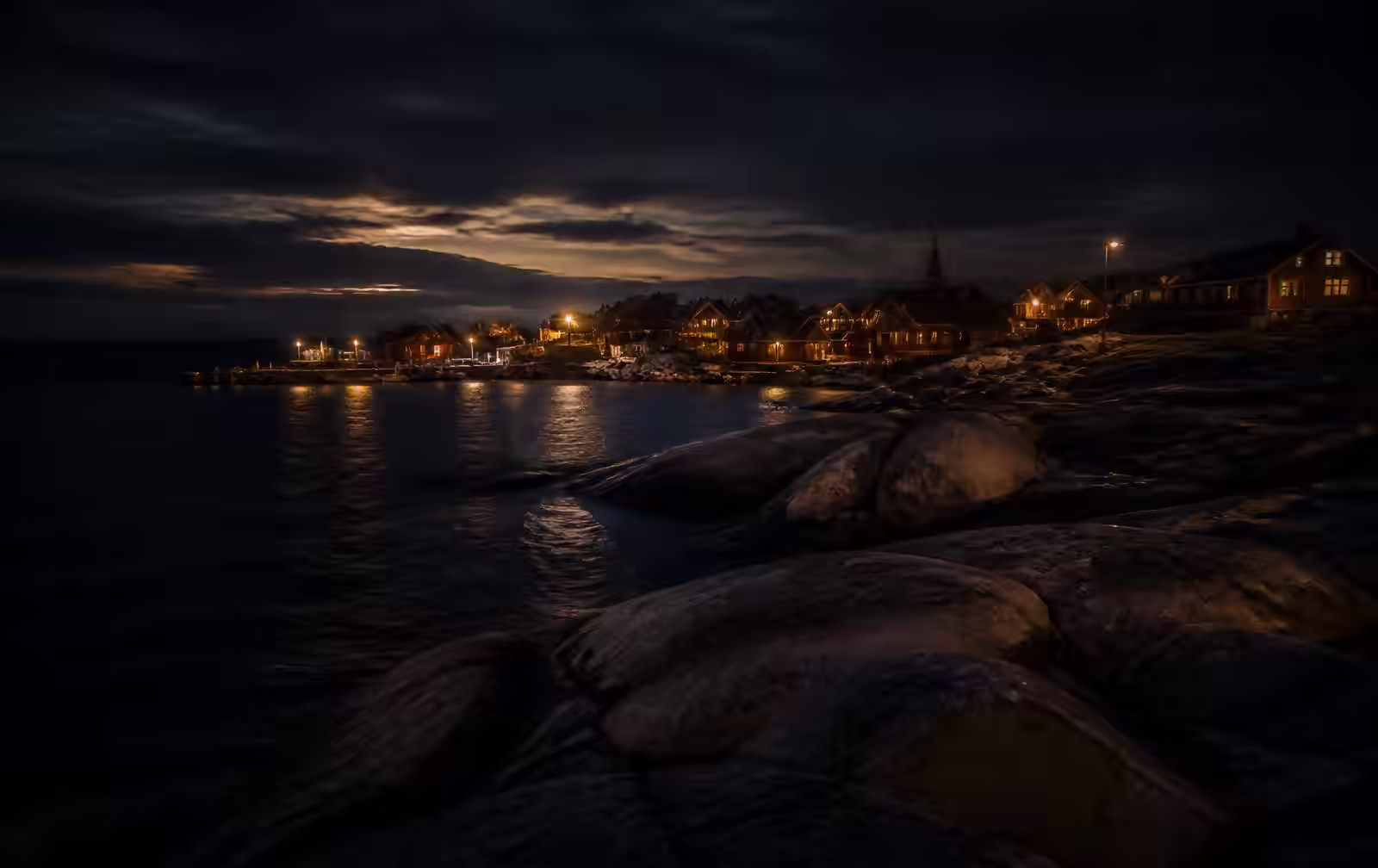 Baltic island coastline at night with granite rocks meeting dark sea and a small harbour village of warm lit windows in the distance