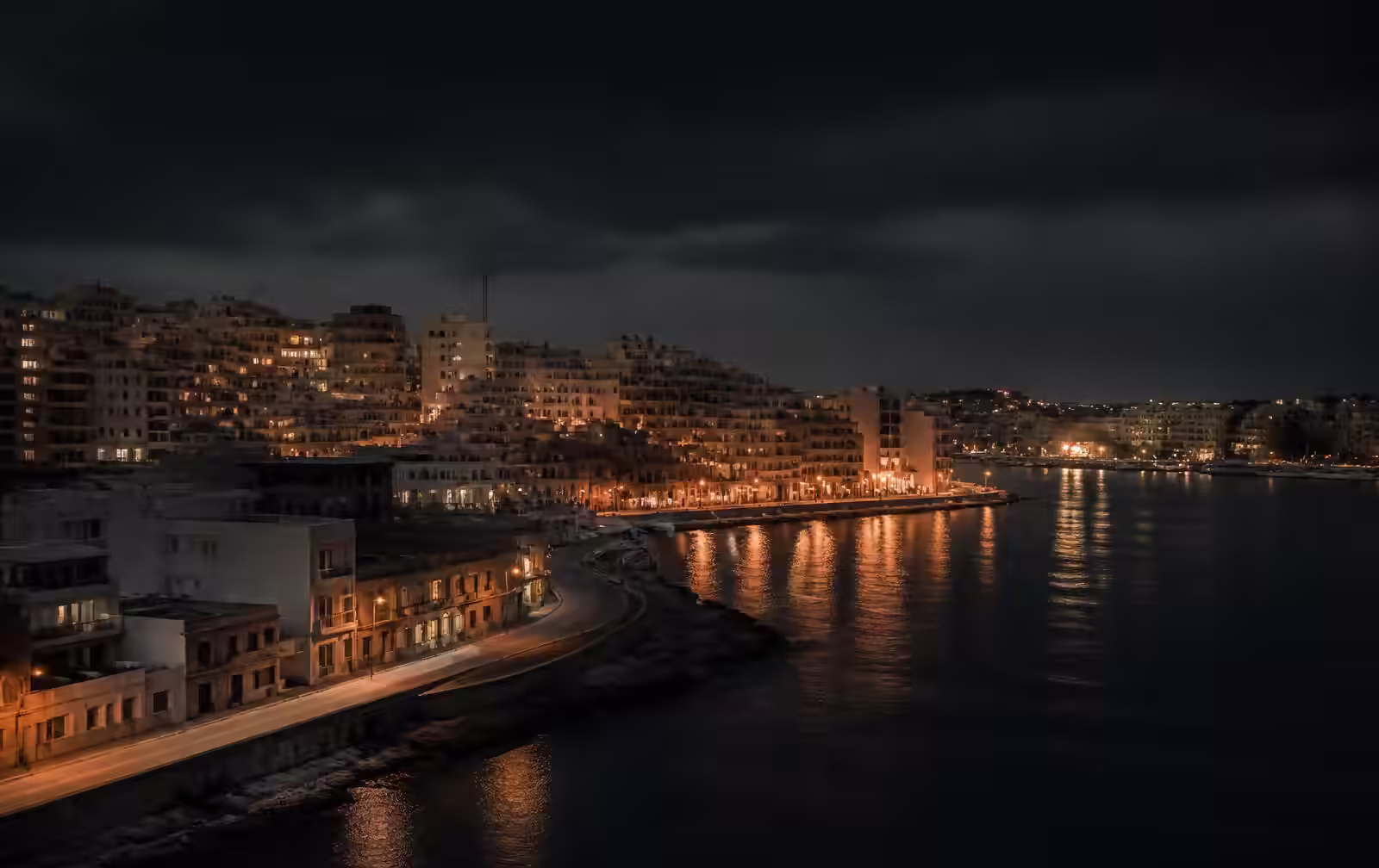 A dense low-rise island city at night, warm amber street lights reflecting across dark harbour water