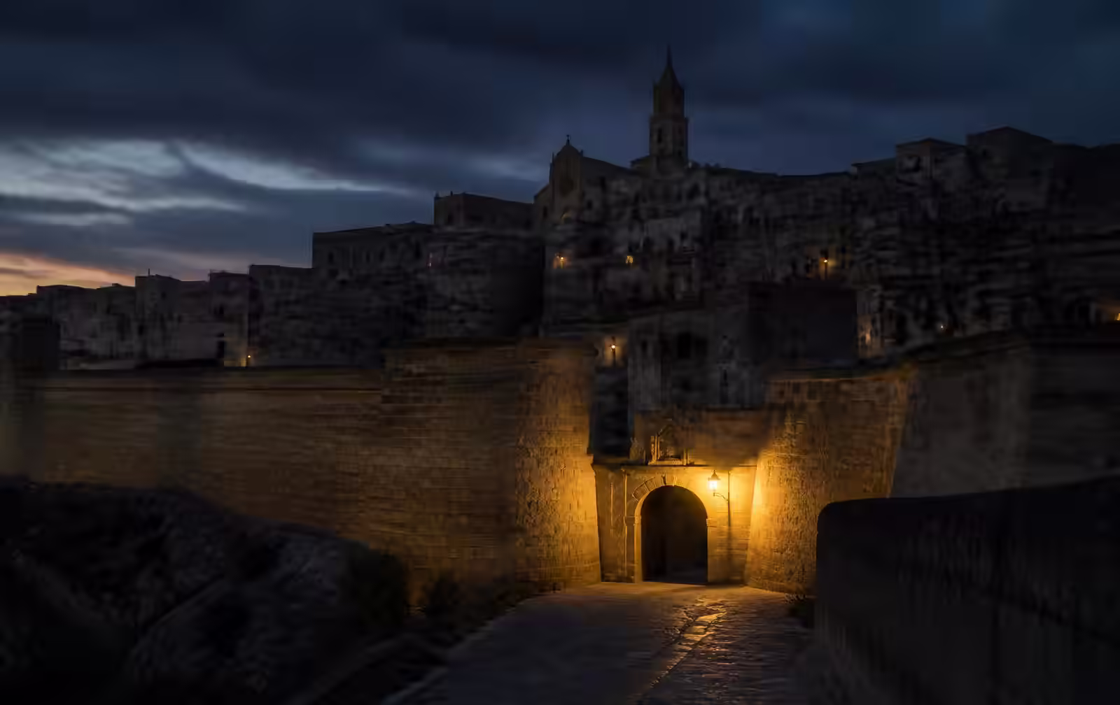 A walled hilltop town at dusk with honey limestone bastions and a single warmly lit arched gateway