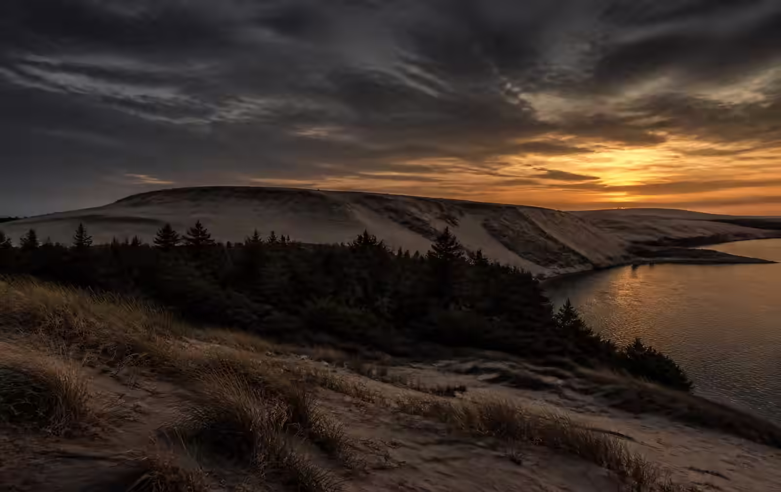 Curonian Spit at dusk, high pale sand dunes above dark pine forest with the still Curonian Lagoon below and a heavy grey and amber sky