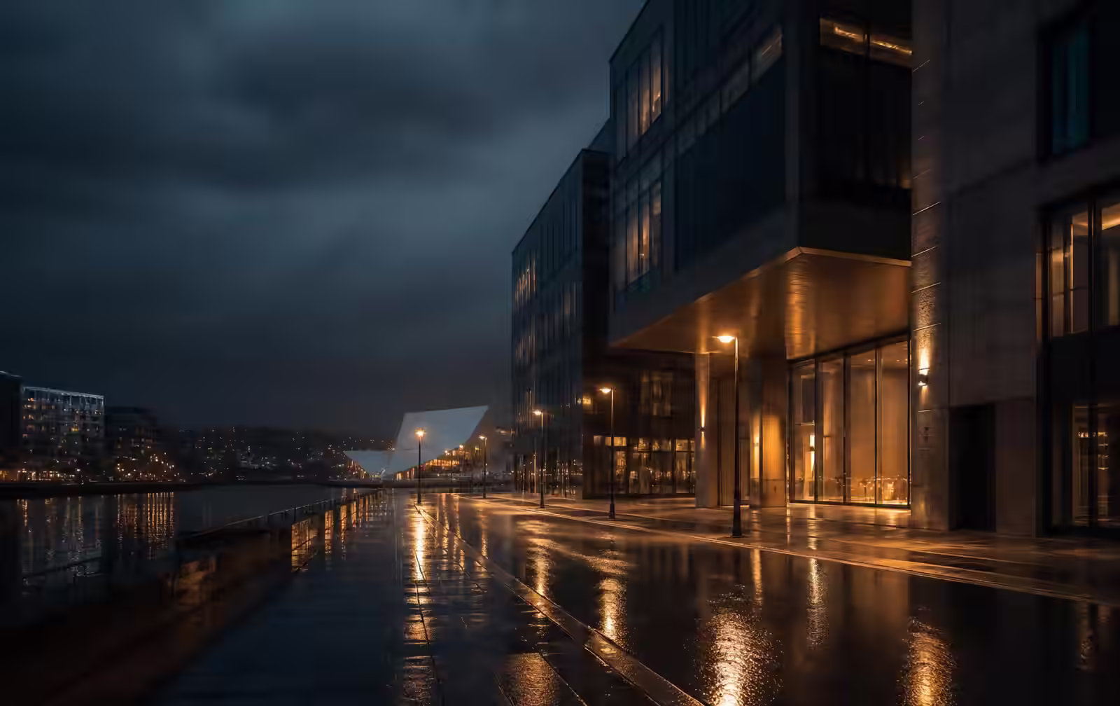 A rain-wet Oslo street at night beside dark modern architecture, warm amber window light reflected in the pavement, harbour water beyond