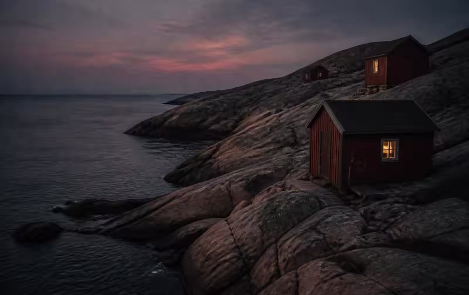 A rugged Swedish granite coastline at dusk, smooth rock slabs above dark calm sea with small red wooden huts showing warm lit windows