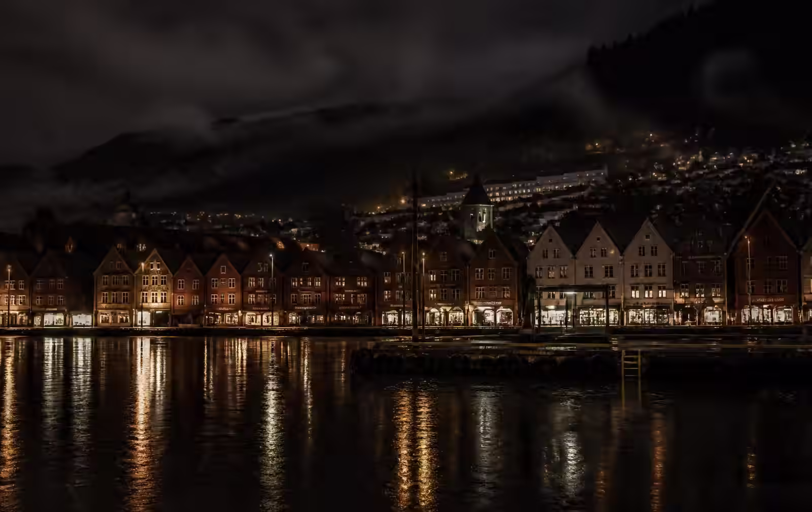 Bergen’s historic wooden harbour buildings at night in the rain, warm lamplight reflected in black water, mist on the hills behind