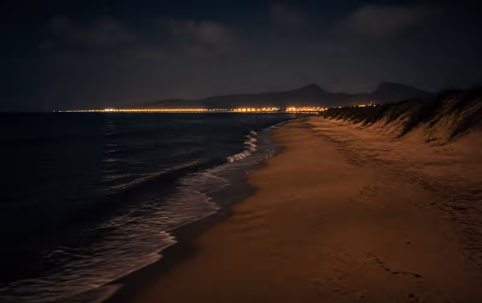 Long empty sandy Mediterranean beach at night with dark surf and distant coastal lights