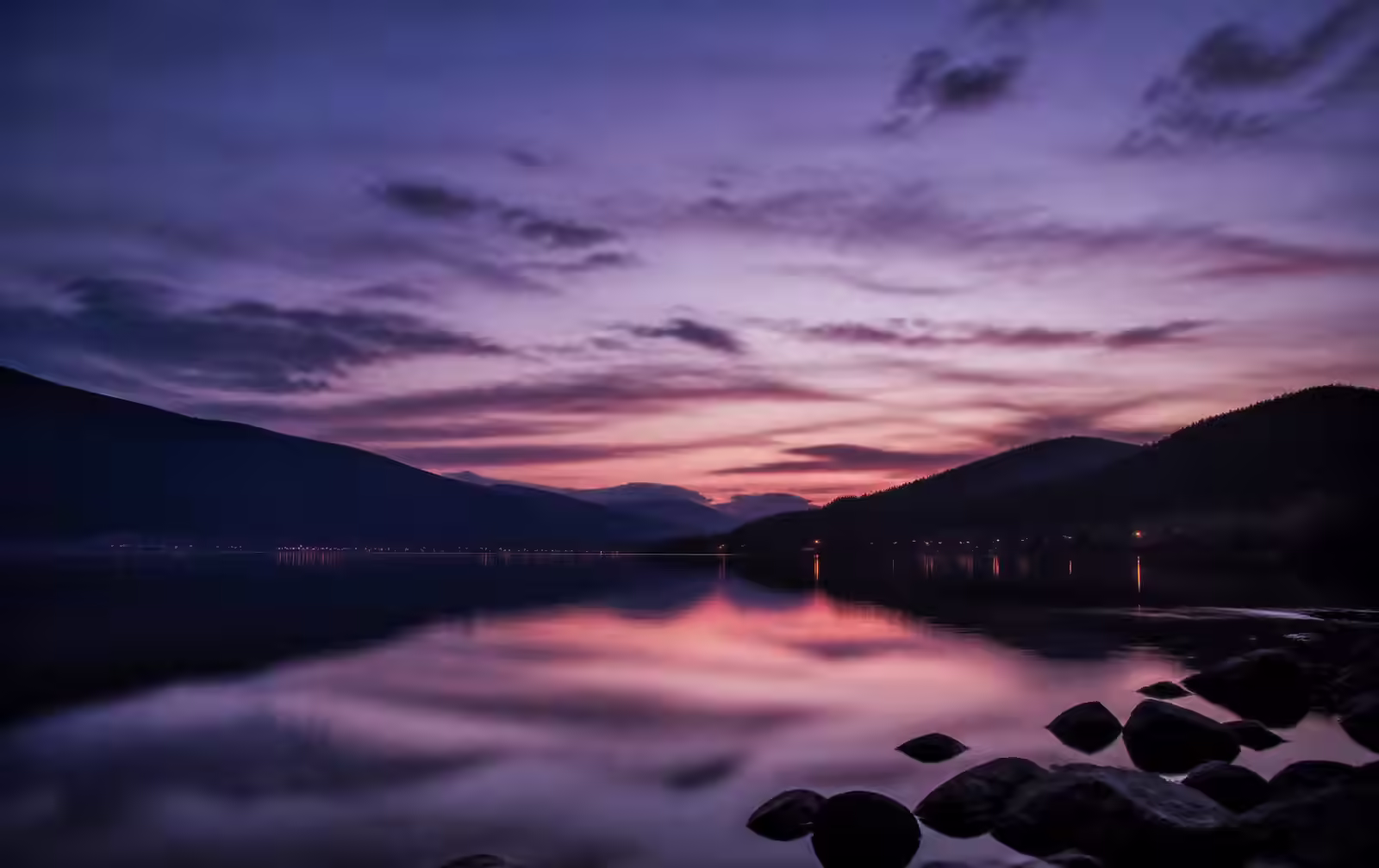 Calm freshwater lake at dusk ringed by low dark hills, with violet light on still water and a few distant shore lights