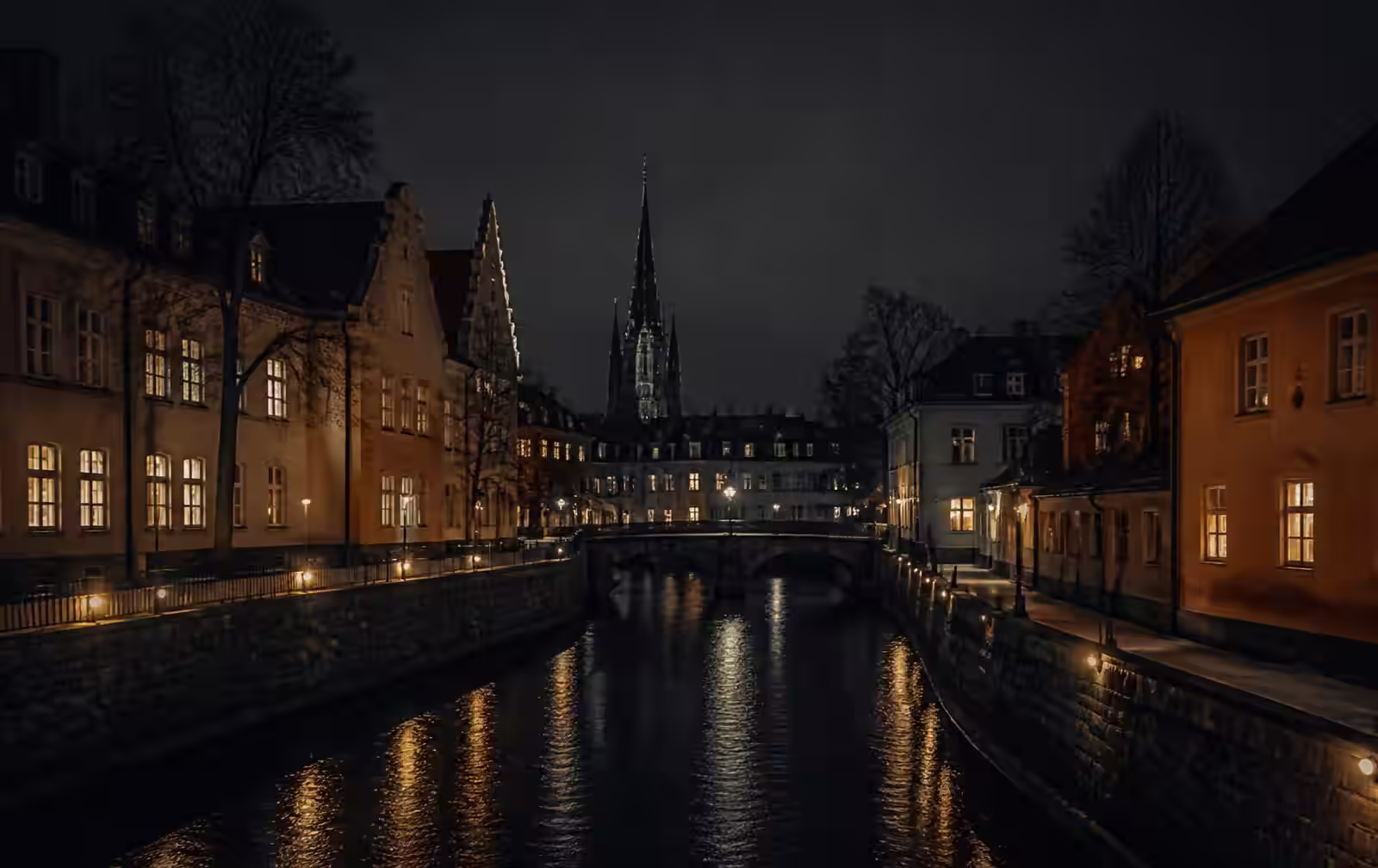 A small historic Swedish city at night, a narrow river between stone embankments with warm lit windows and a cathedral spire in the dark distance