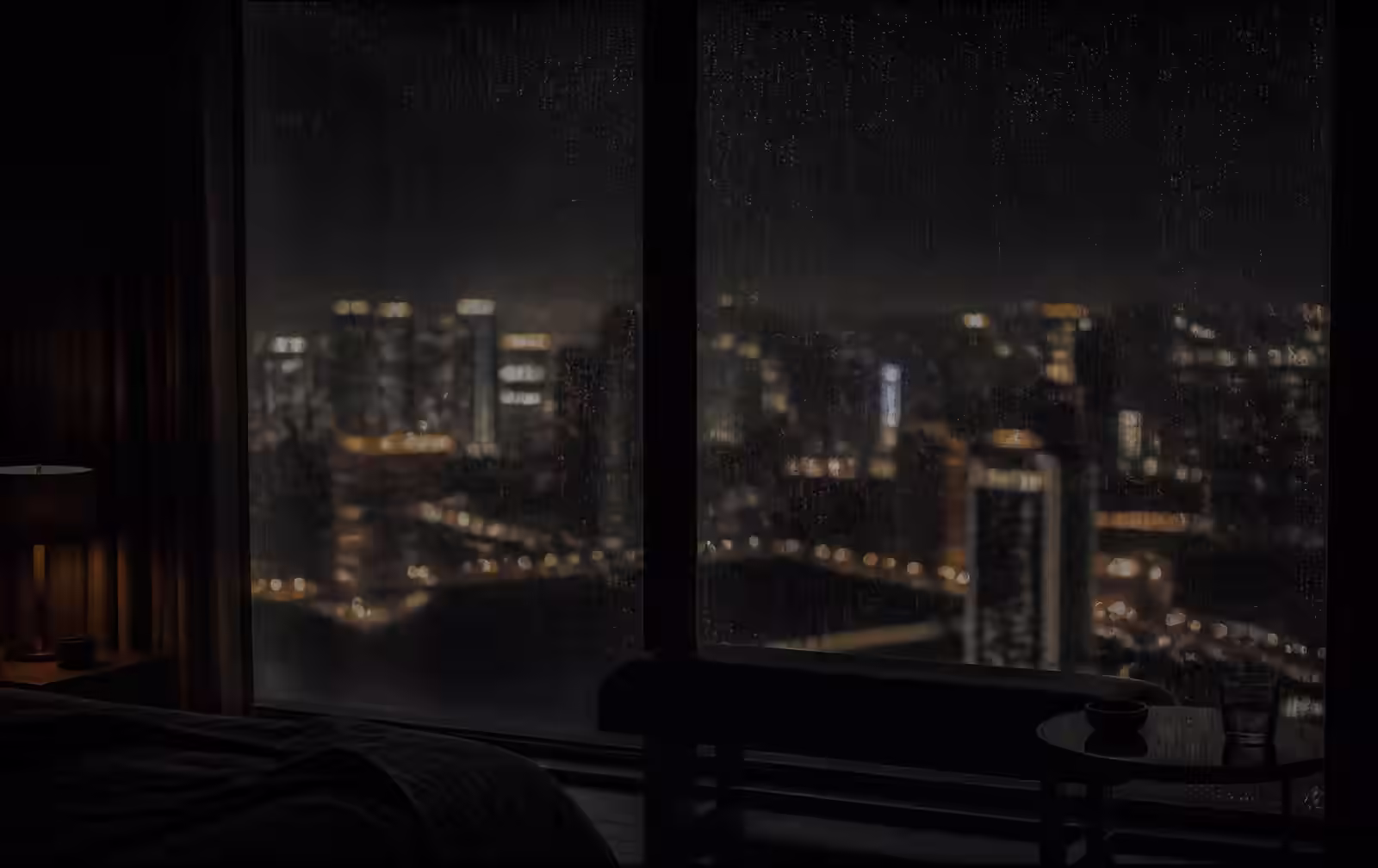 A rain-streaked window of a dark hotel suite overlooking distant city lights at night