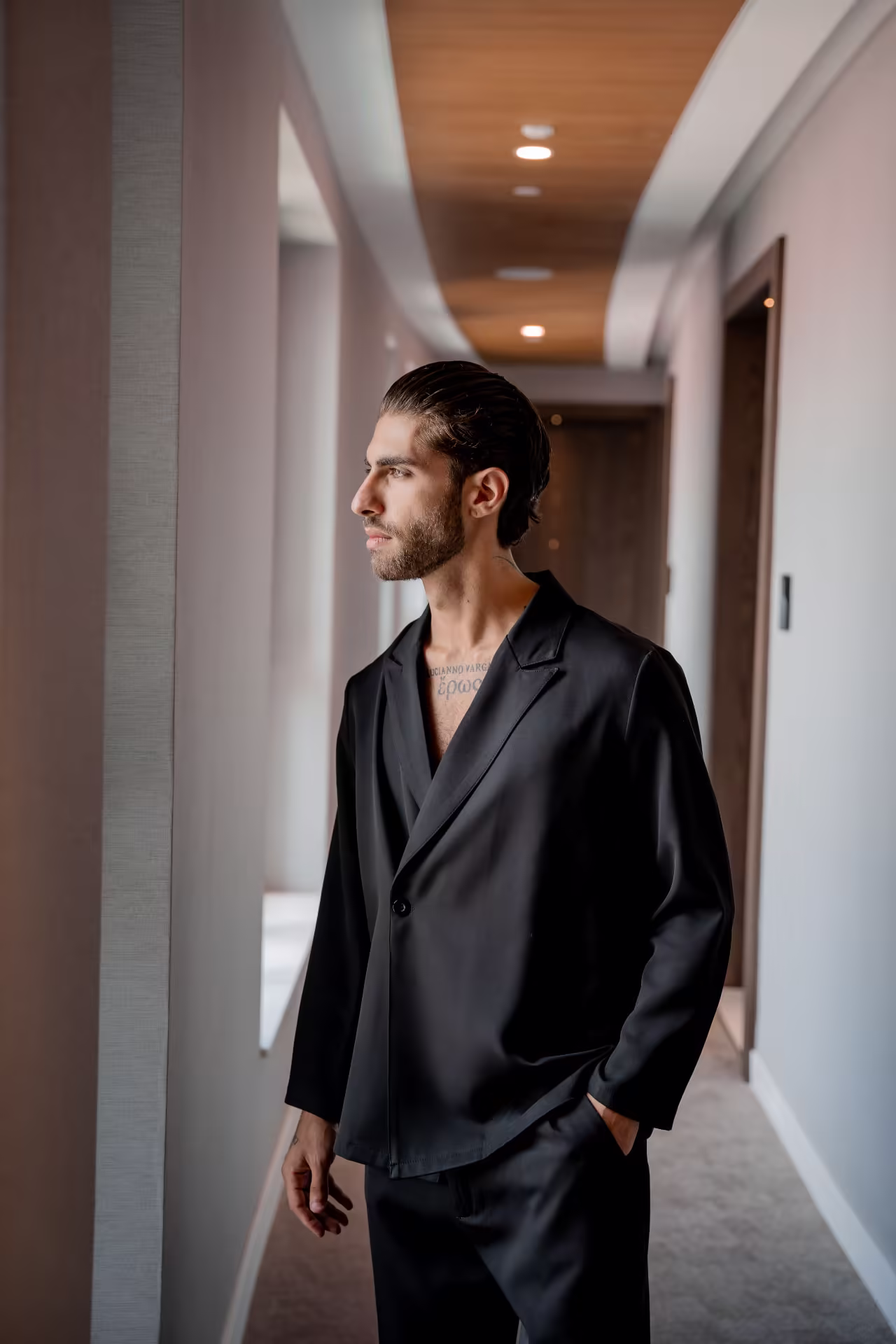 Lucianno Vargas in profile in a hotel corridor, black blazer, one hand in his pocket