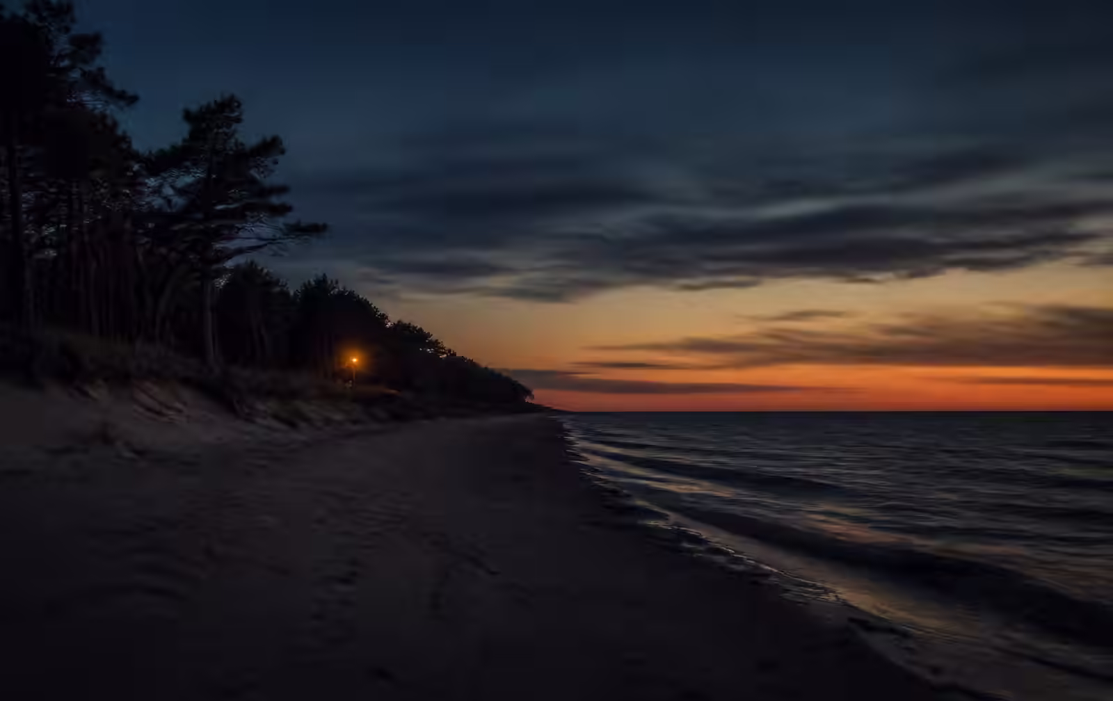 Baltic Sea beach near Palanga at dusk, empty pale sand, dark calm water, pine trees along the dune line and a single distant lamp