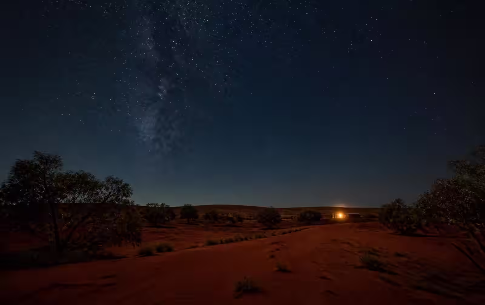 Remote red desert country at night under a vast star field with one distant warm light from a small building