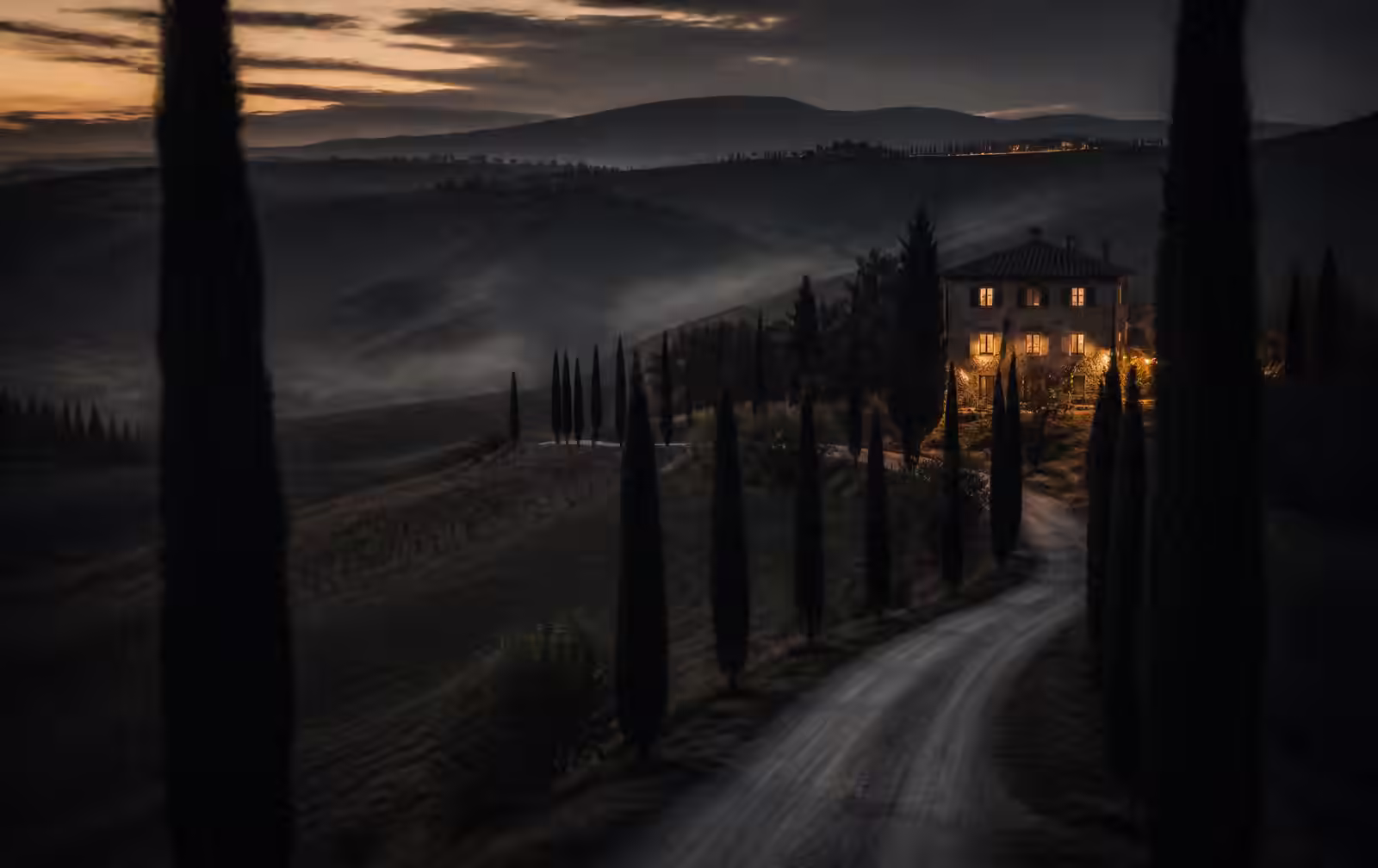 A cypress-lined road through misty Val d’Orcia hills at dusk leading to a stone villa with lit windows
