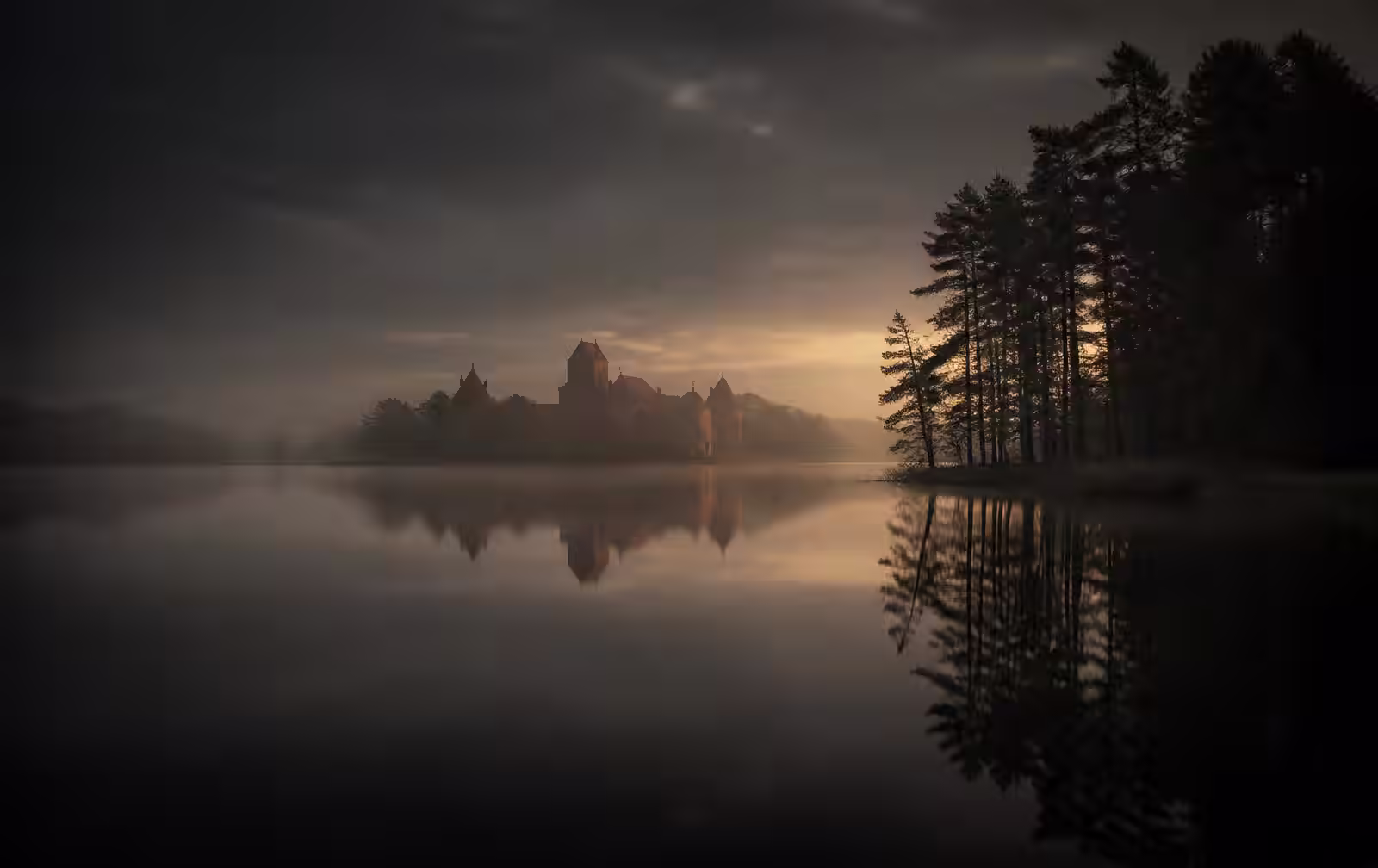 Still lake at Trakai in the evening mist, dark mirrored water, a distant red brick island castle silhouette and pine trees along the shore