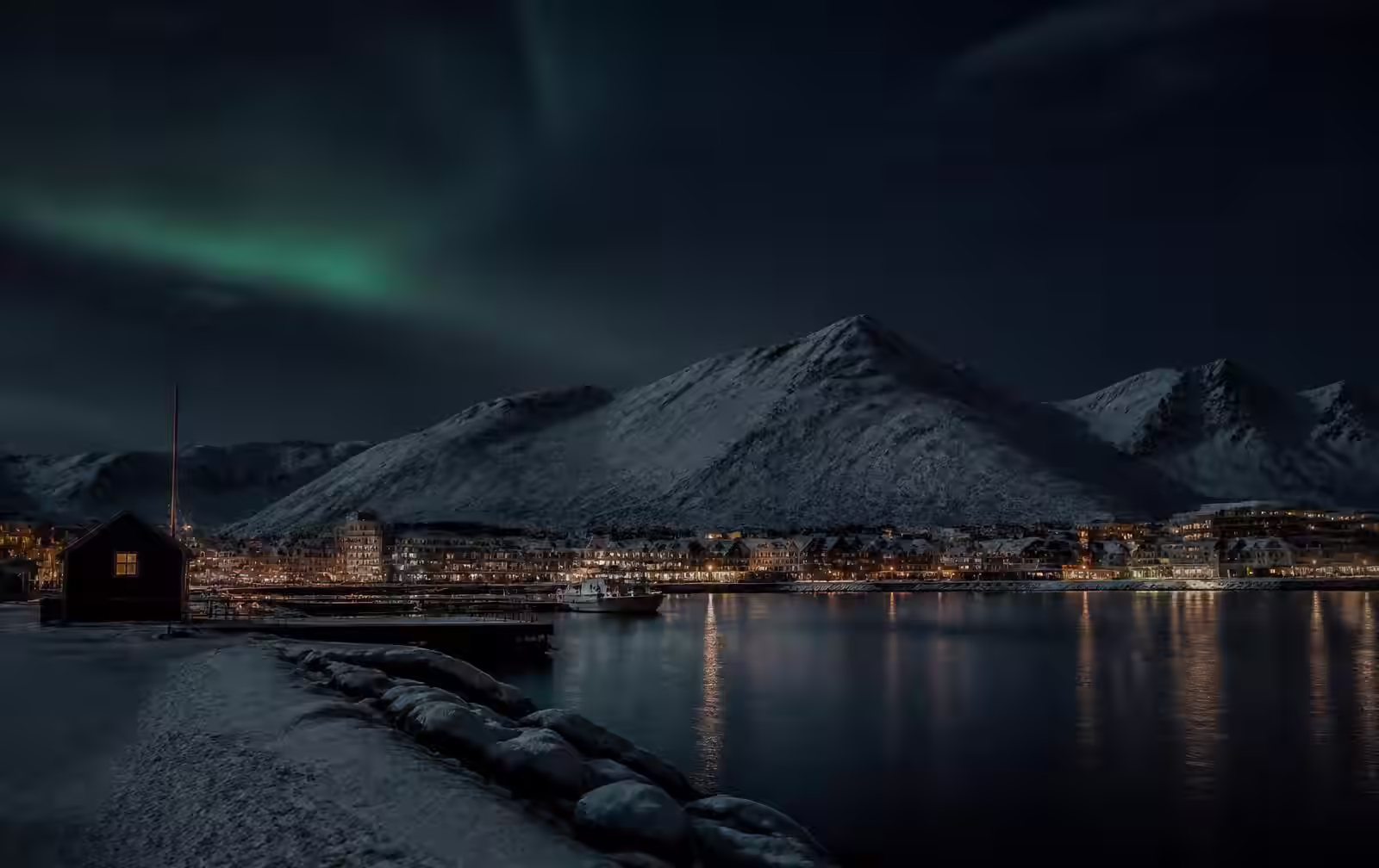 An Arctic Norwegian city at polar night, snow-covered harbour and dark mountains across the water, warm gold windows and a faint green aurora high above