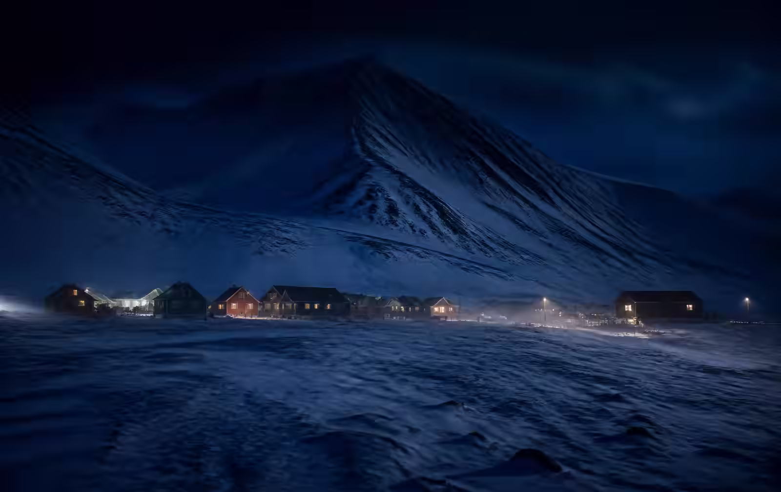 A small high-Arctic settlement during polar night, coloured wooden buildings with warm windows on a snow plain beneath dark glaciated mountains