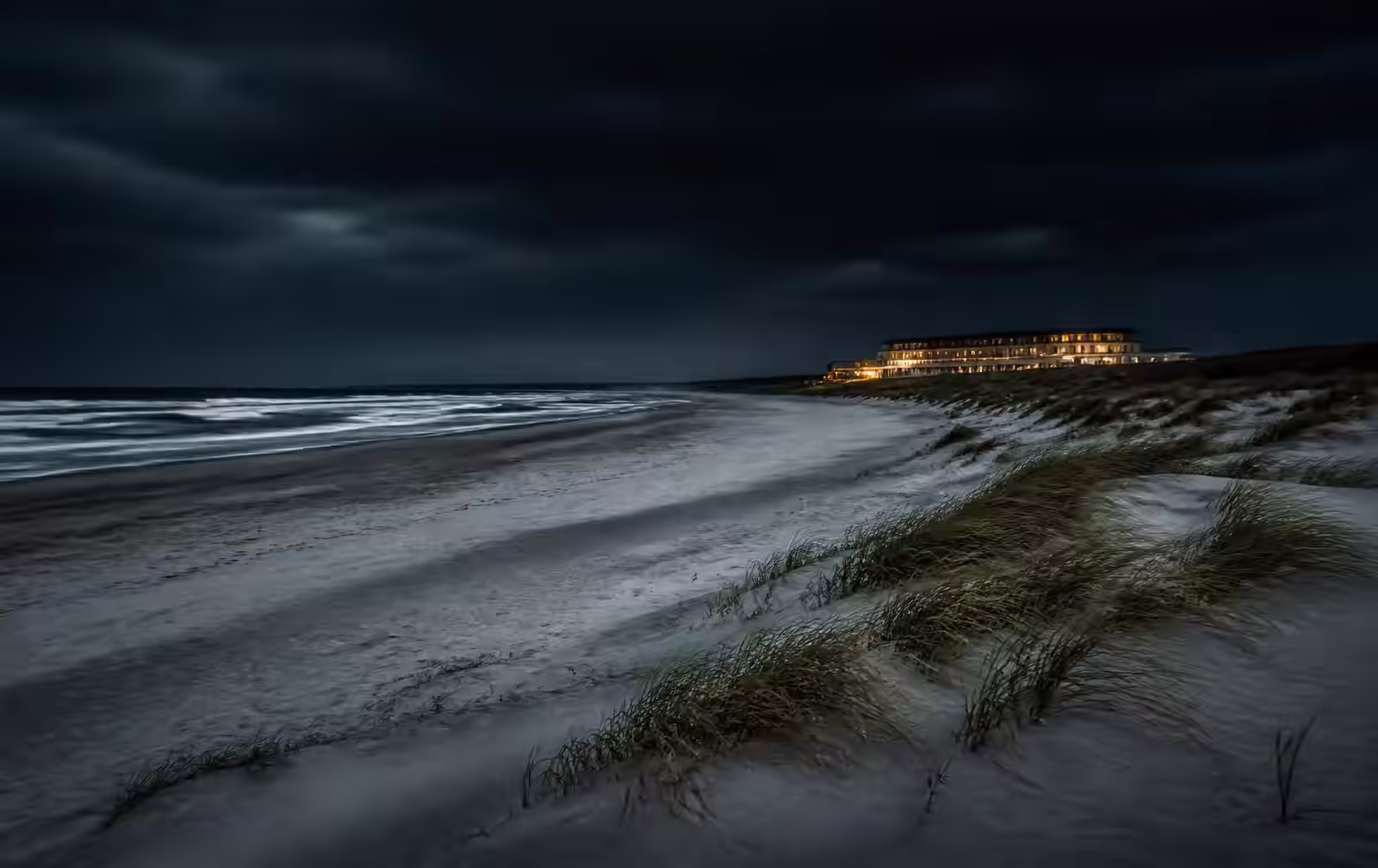 Empty North Sea beach at night with dune grass, dark surf and a distant warmly lit beachfront hotel