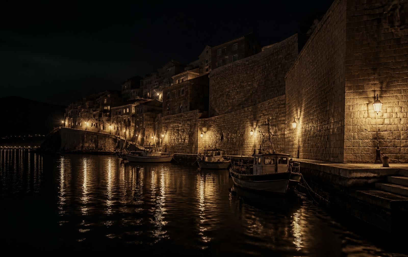 Old stone-walled harbour town at night with lamplit sea walls, moored fishing boats and dark water