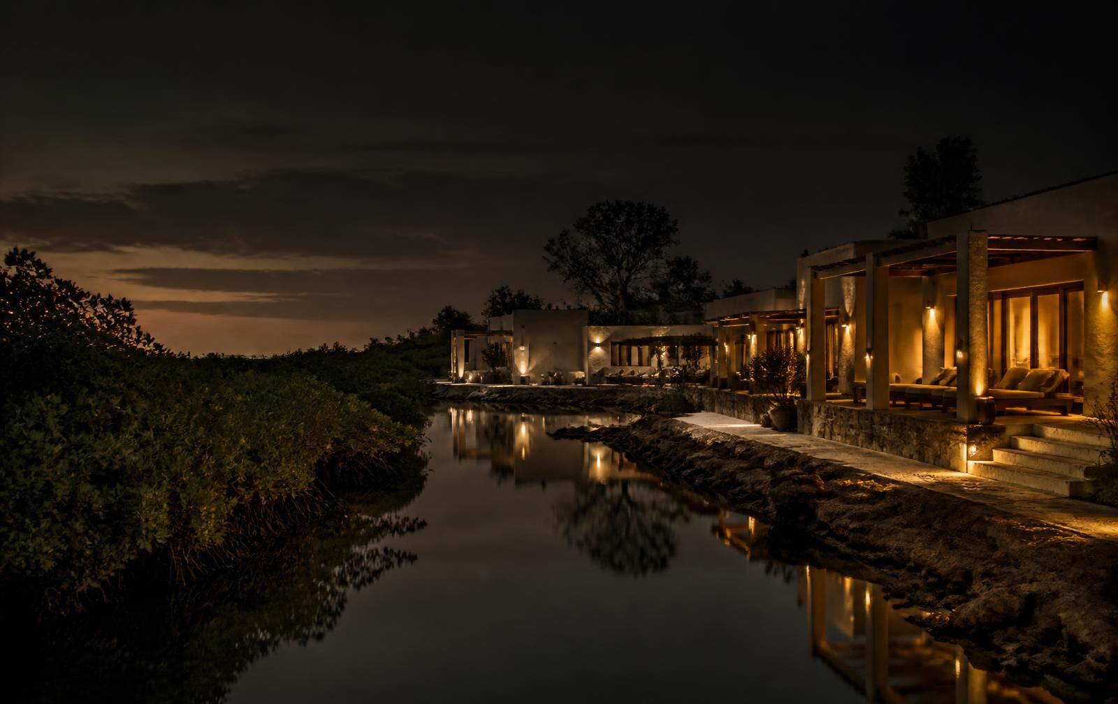 A quiet coastal resort beside mangroves and a still tidal creek at dusk with warm gold lamplight