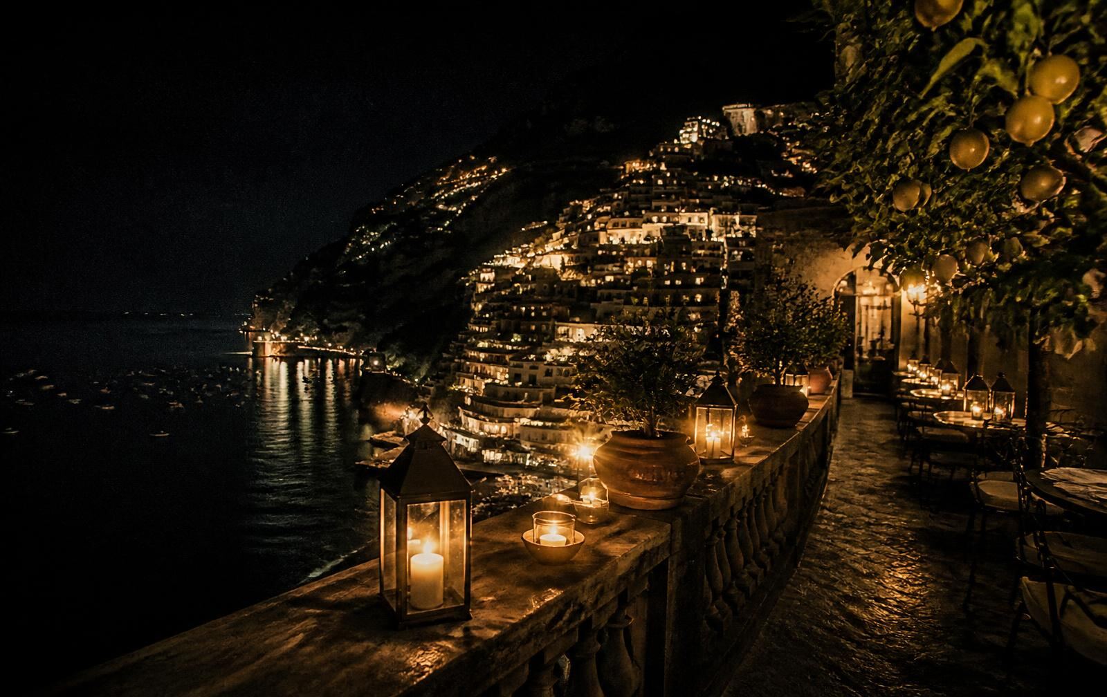 A candlelit Amalfi Coast terrace at night with lemon trees above cliffside village lights and dark sea