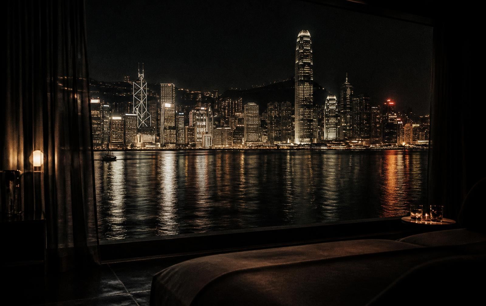 A harbour city at night seen from a darkened private interior, with warm gold lights reflected on black water