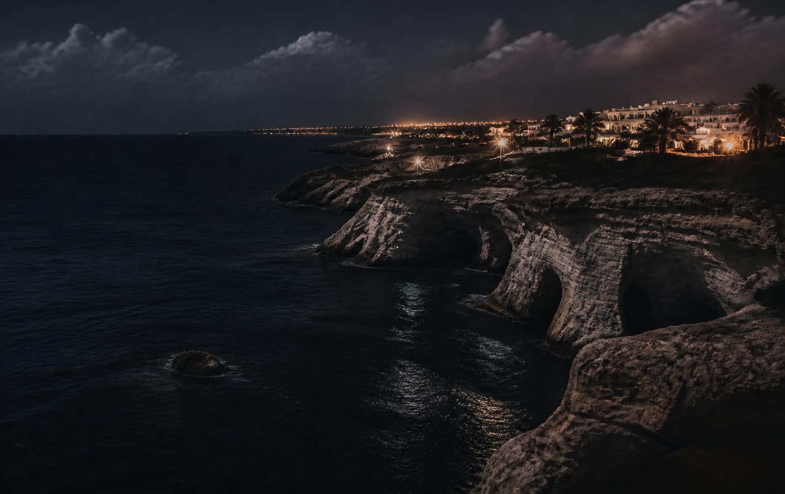 South-east Cyprus limestone sea caves at night with dark water and distant warm resort lights