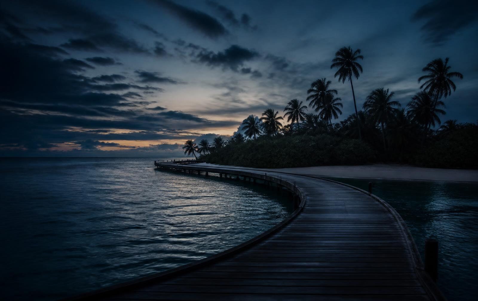 A long wooden jetty curving over deep blue water toward a palm island at twilight