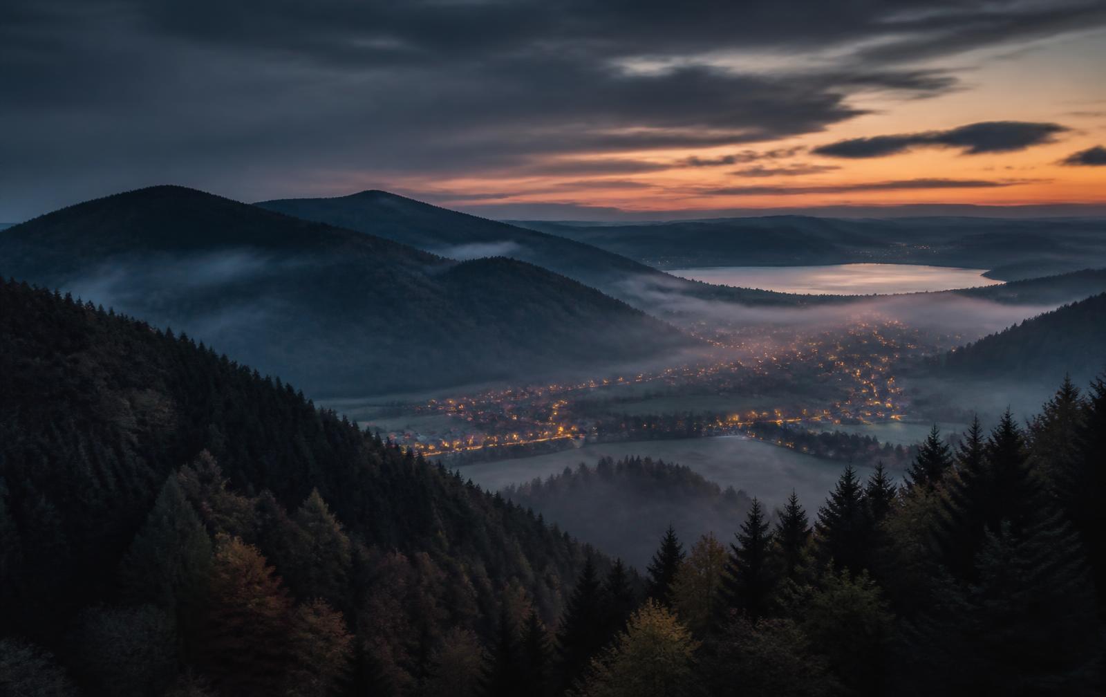 Dark forested hills above a misty valley at dusk in south-west Germany, with village lights and a distant lake
