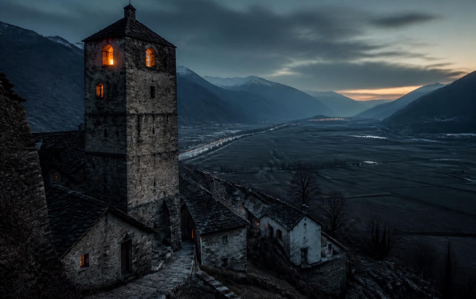 An old stone tower above village rooftops at dusk with a wide alpine valley and distant mountains