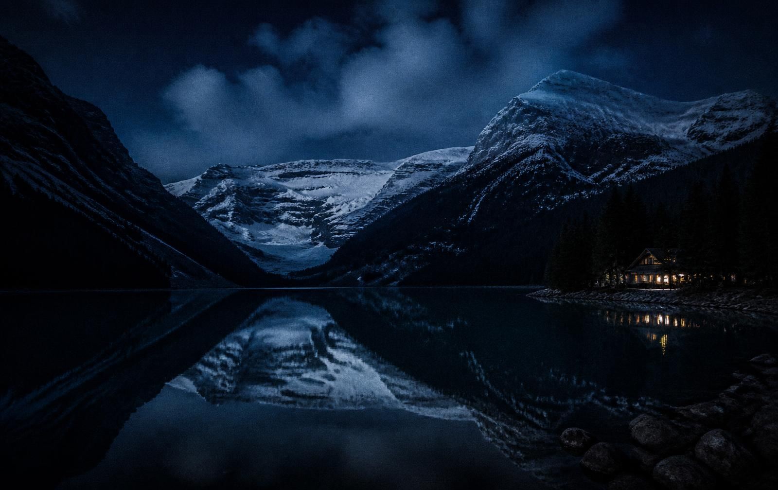 Still dark glacial lake at night reflecting snow-covered peaks with one warm lodge light at the shore