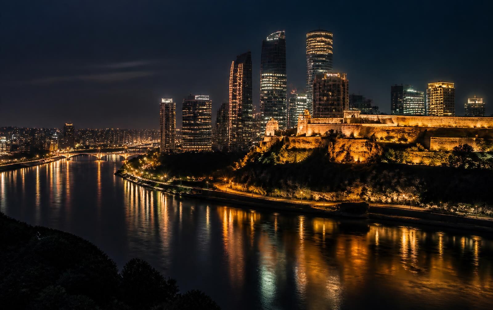 A riverside capital skyline at night with lit towers, a floodlit fortress bluff and gold reflections on dark water