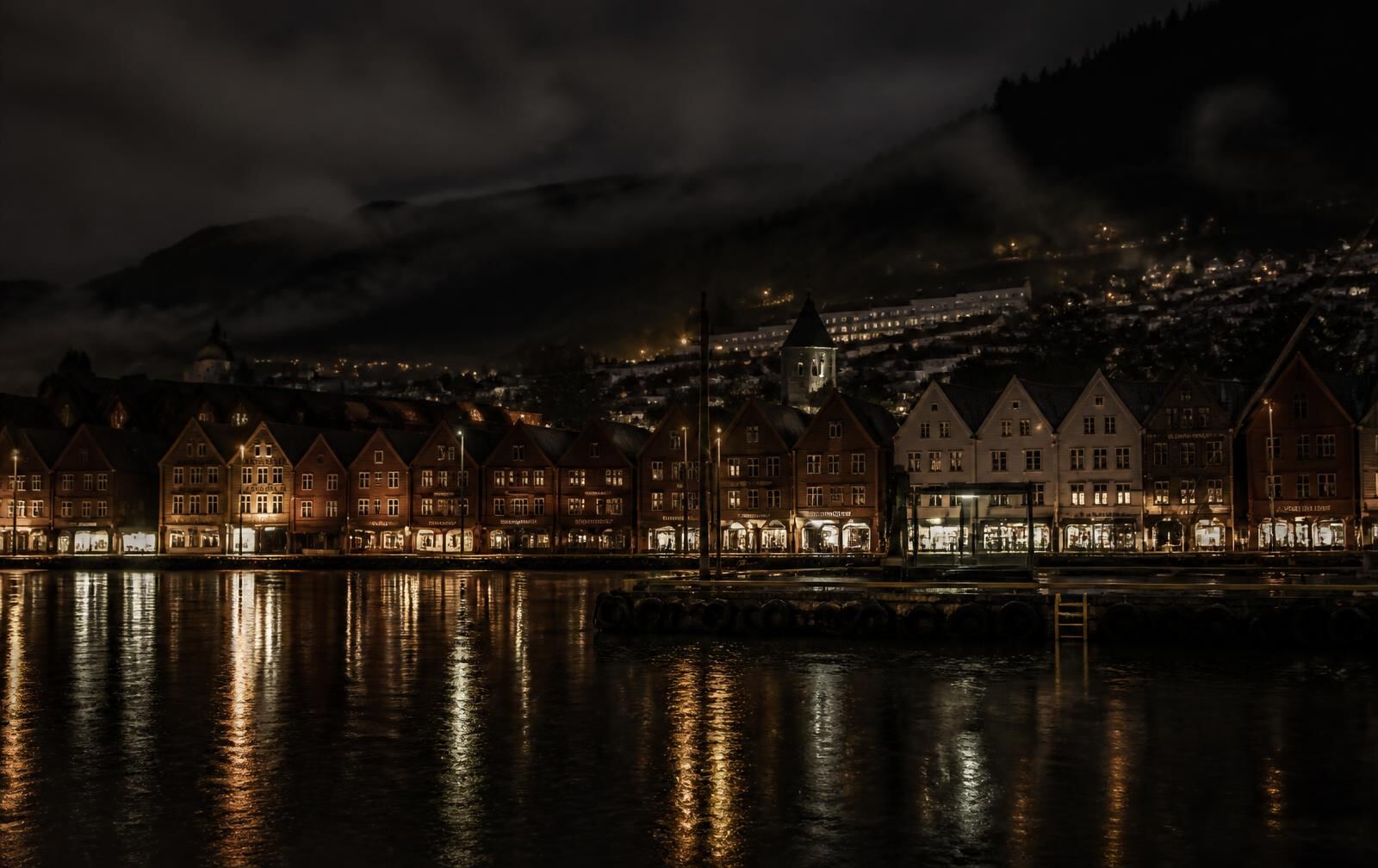 Bergen’s historic wooden harbour buildings at night in the rain, warm lamplight reflected in black water, mist on the hills behind
