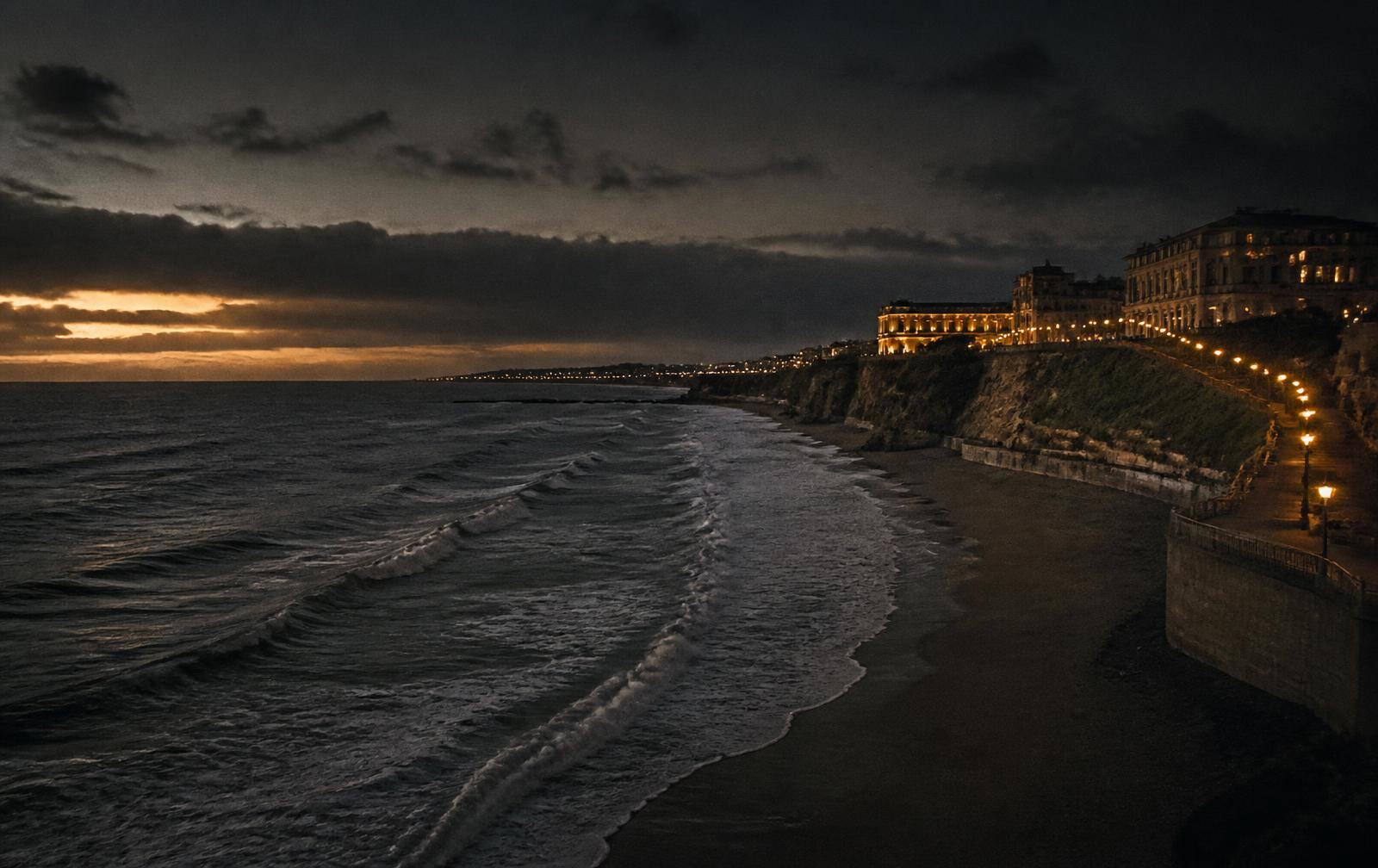 The Atlantic coast at Biarritz at dusk with breaking waves below a lamplit clifftop promenade