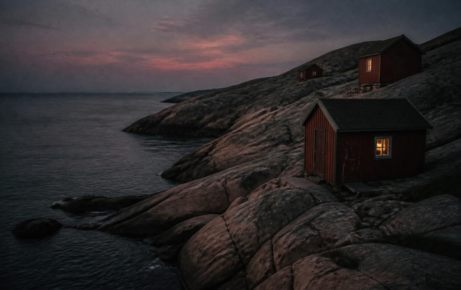 A rugged Swedish granite coastline at dusk, smooth rock slabs above dark calm sea with small red wooden huts showing warm lit windows