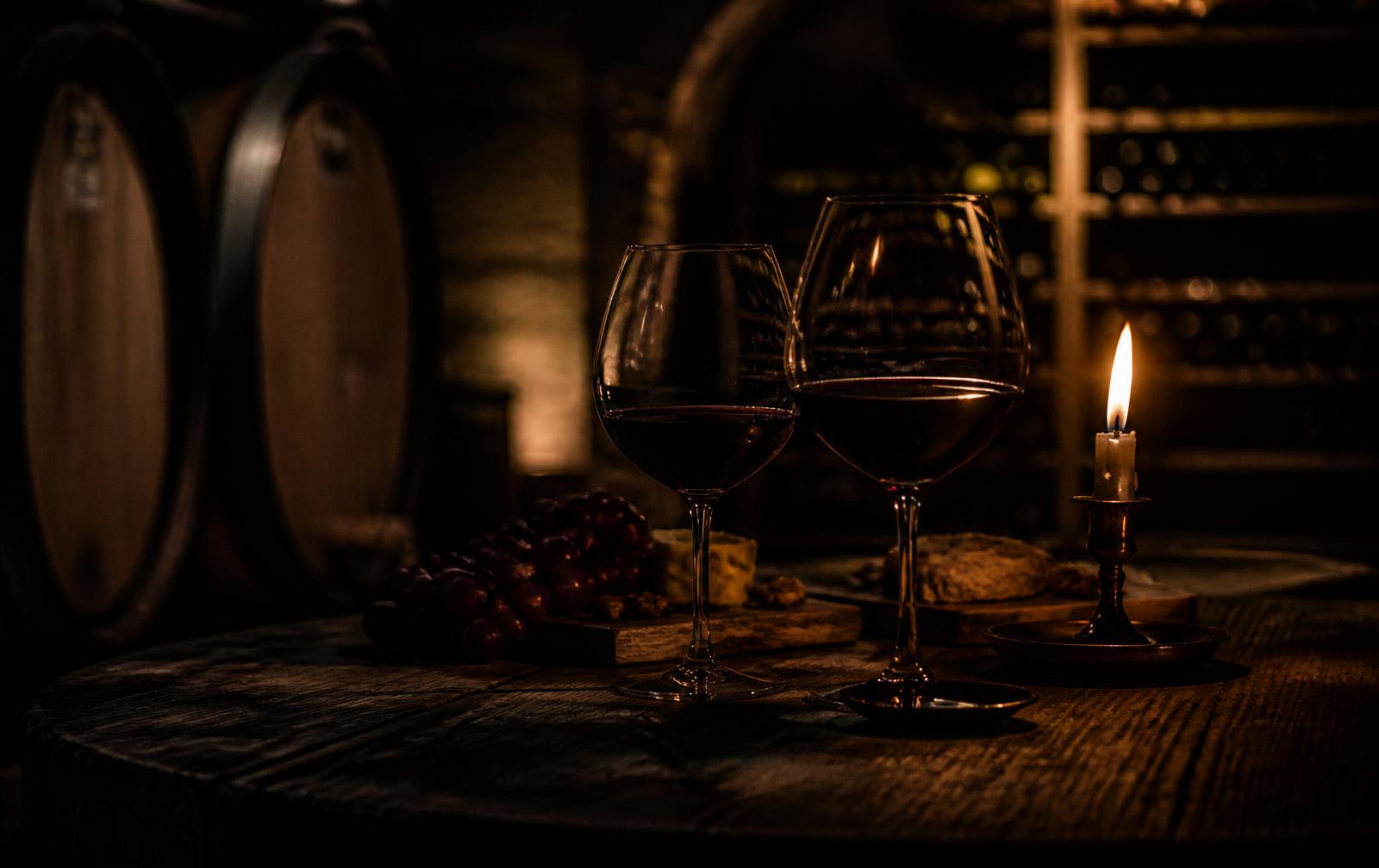 A candlelit table with two glasses of red wine in a dark Bordeaux wine cellar with oak barrels
