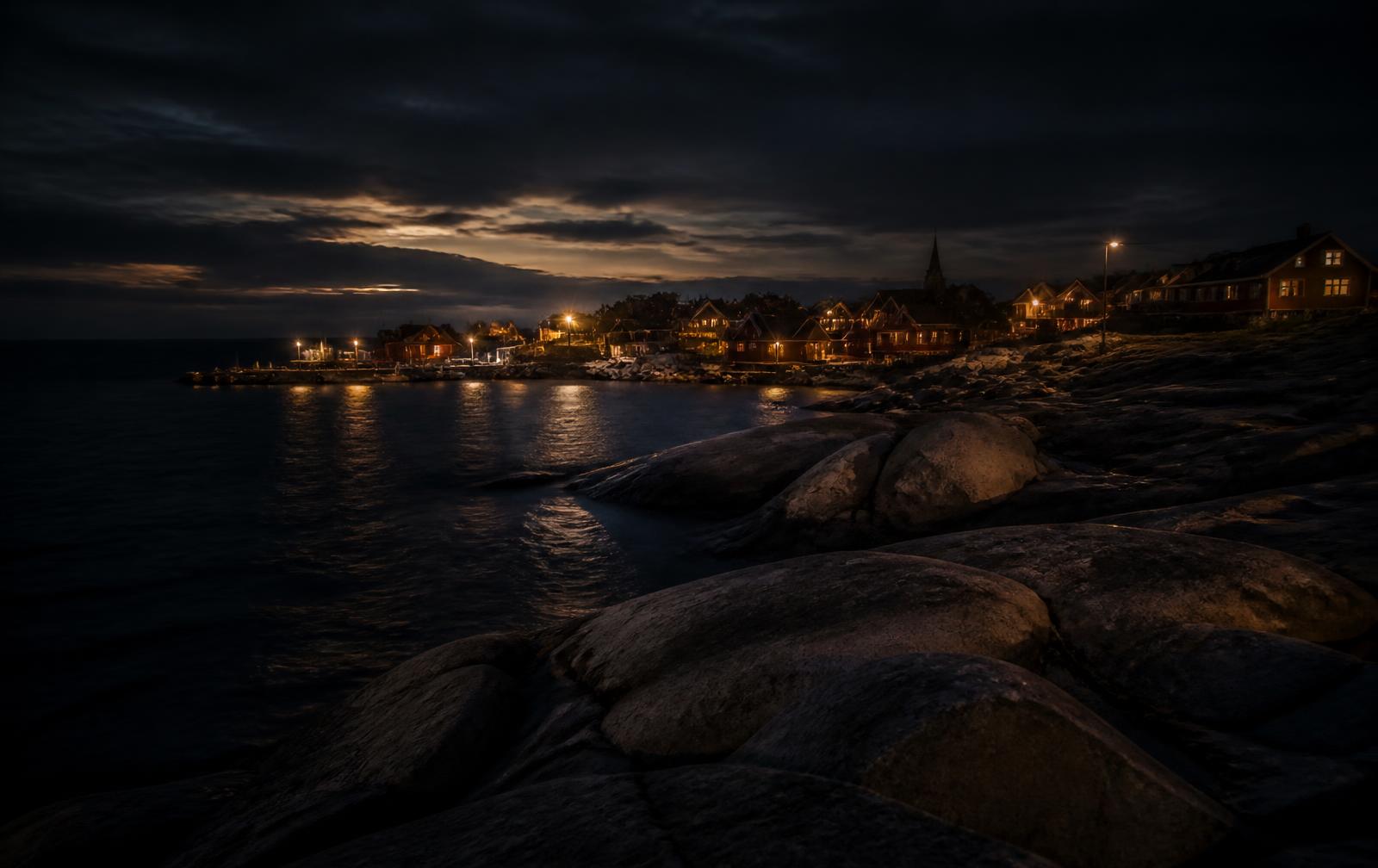 Baltic island coastline at night with granite rocks meeting dark sea and a small harbour village of warm lit windows in the distance