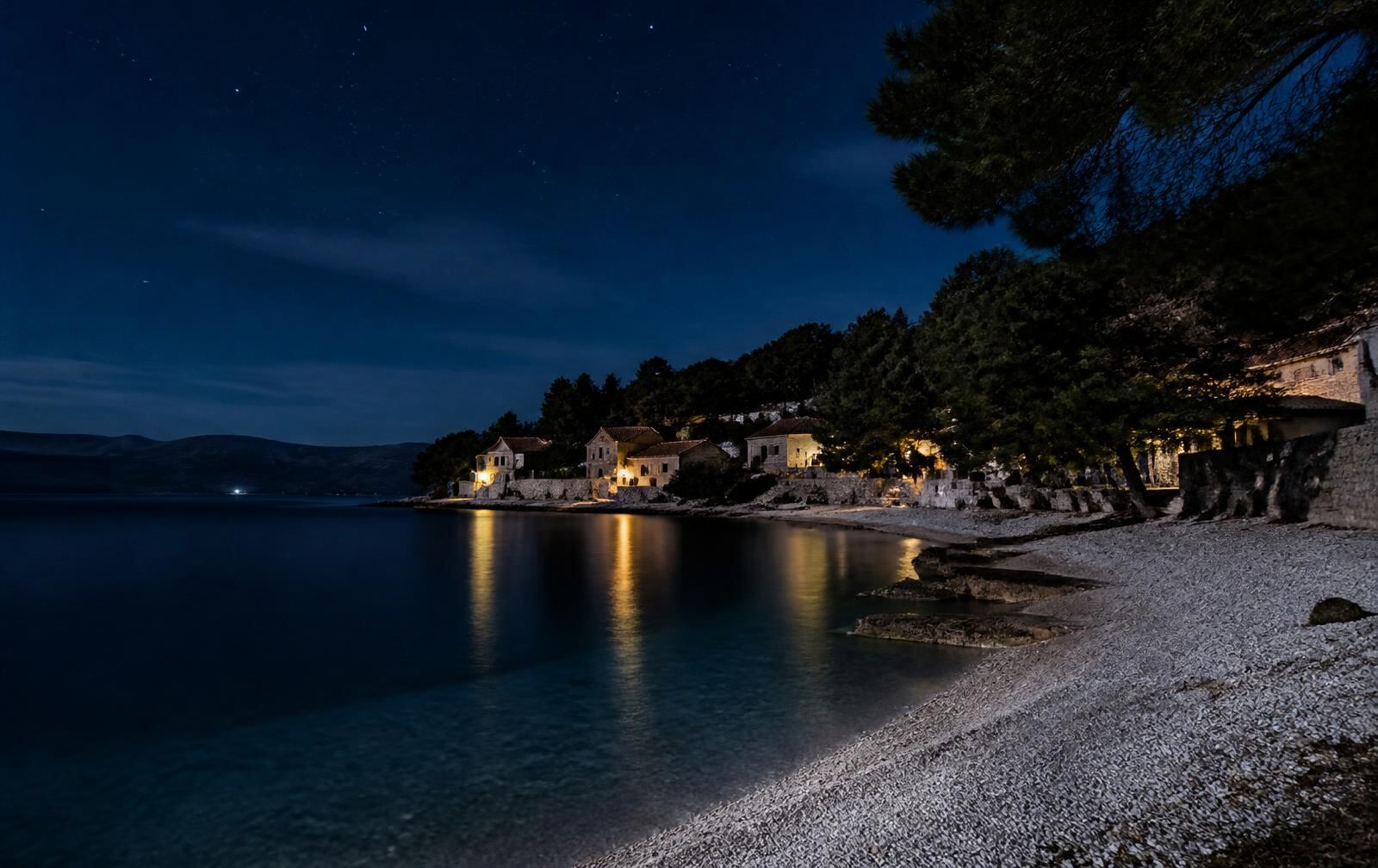 A quiet Croatian island cove at night — pale pebble shore, warm village lights, pine silhouettes under a starlit sky