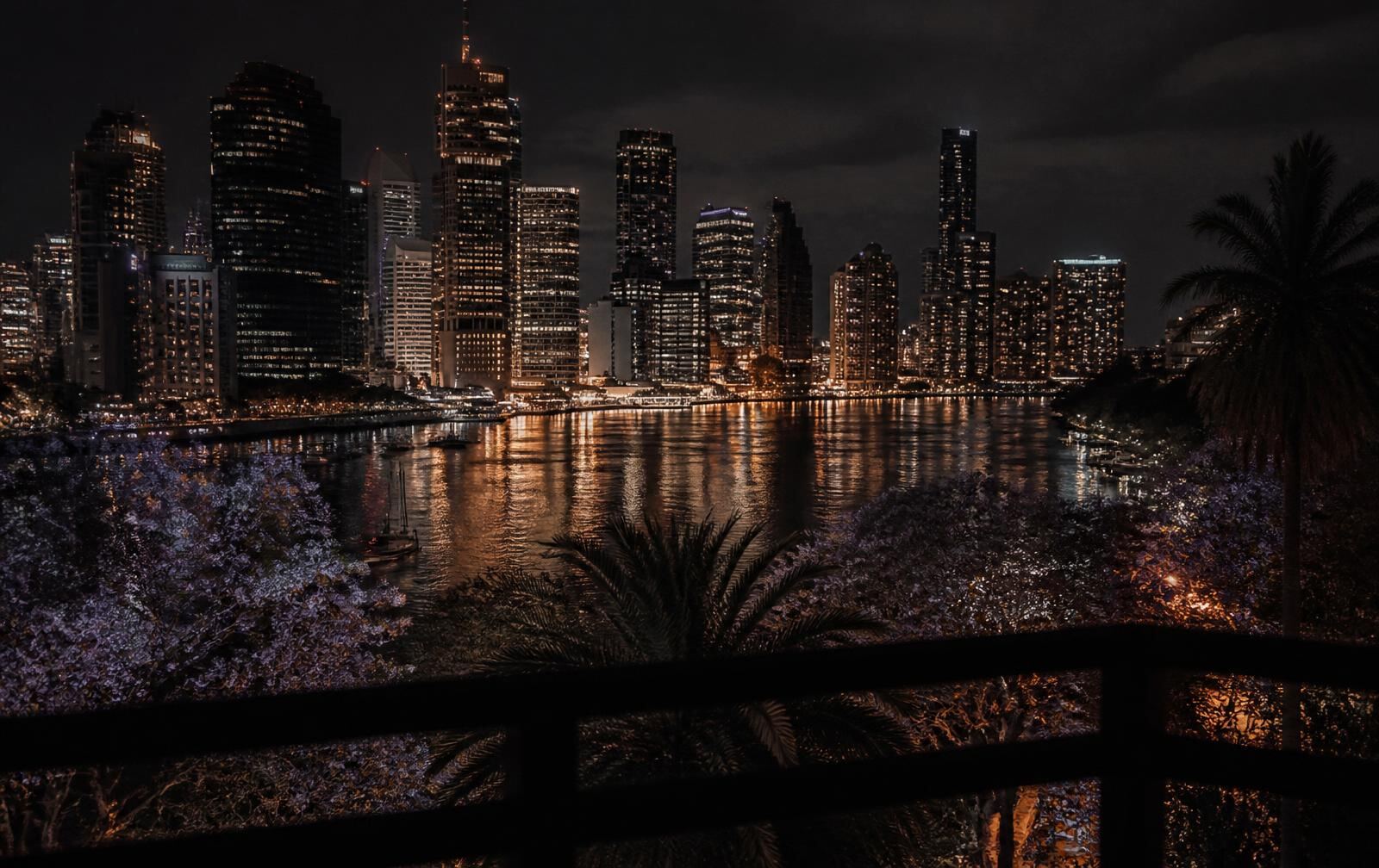 Brisbane's river and glass towers at night with gold reflections on dark water seen past a balcony rail and jacarandas
