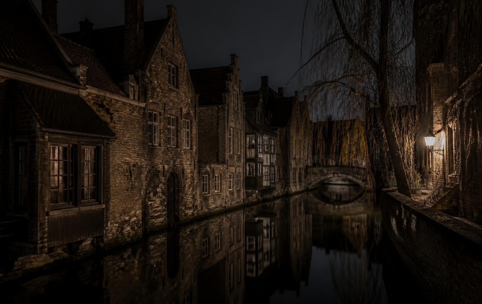 Narrow Bruges canal at night with medieval brick houses, an arched bridge and a single lantern reflected in still water