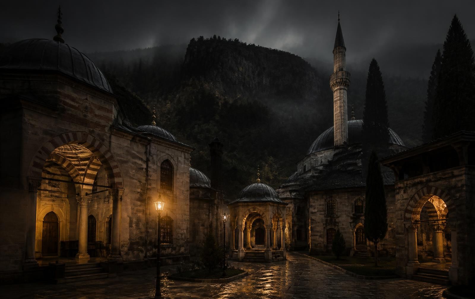 Historic Ottoman-era stone courtyard at night in soft mist with warm lamplight and a forested mountain behind