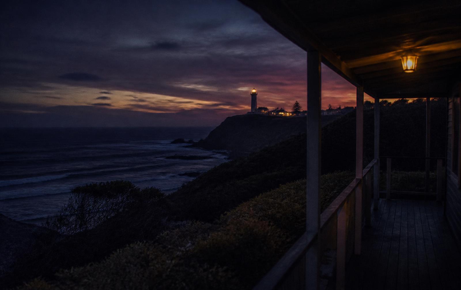 A dark subtropical coastline at dusk with a distant lighthouse headland seen from a lamplit timber verandah