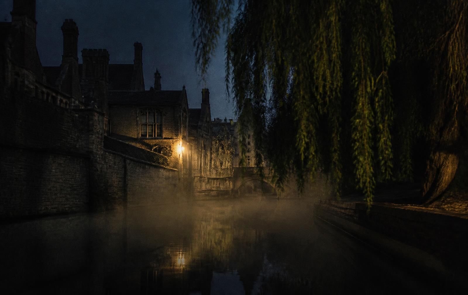 Misty river at night beside old English college stone walls with willows and one warm light
