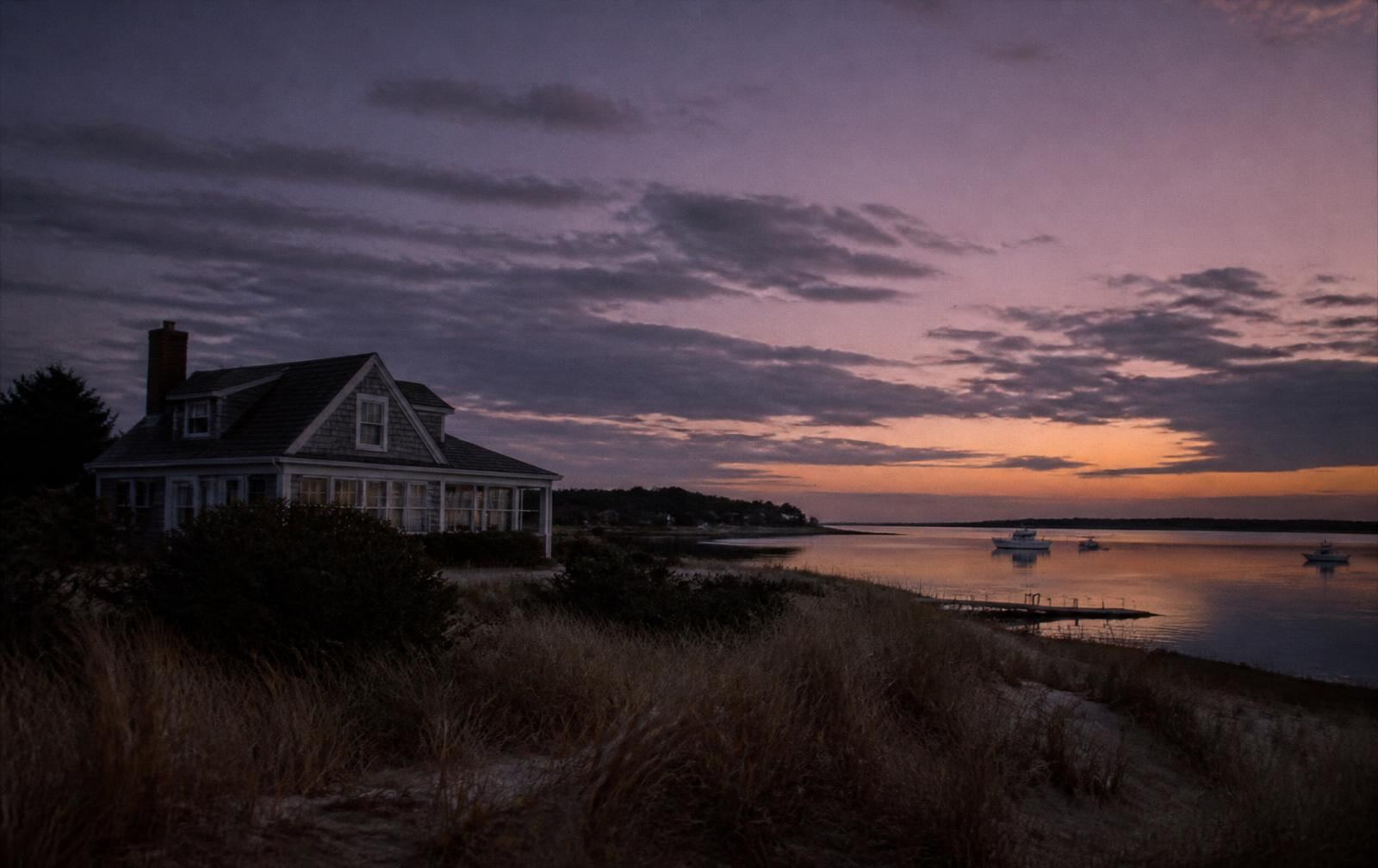A weathered Cape Cod cottage with soft lamplight at dusk, fog settling over a still, empty road