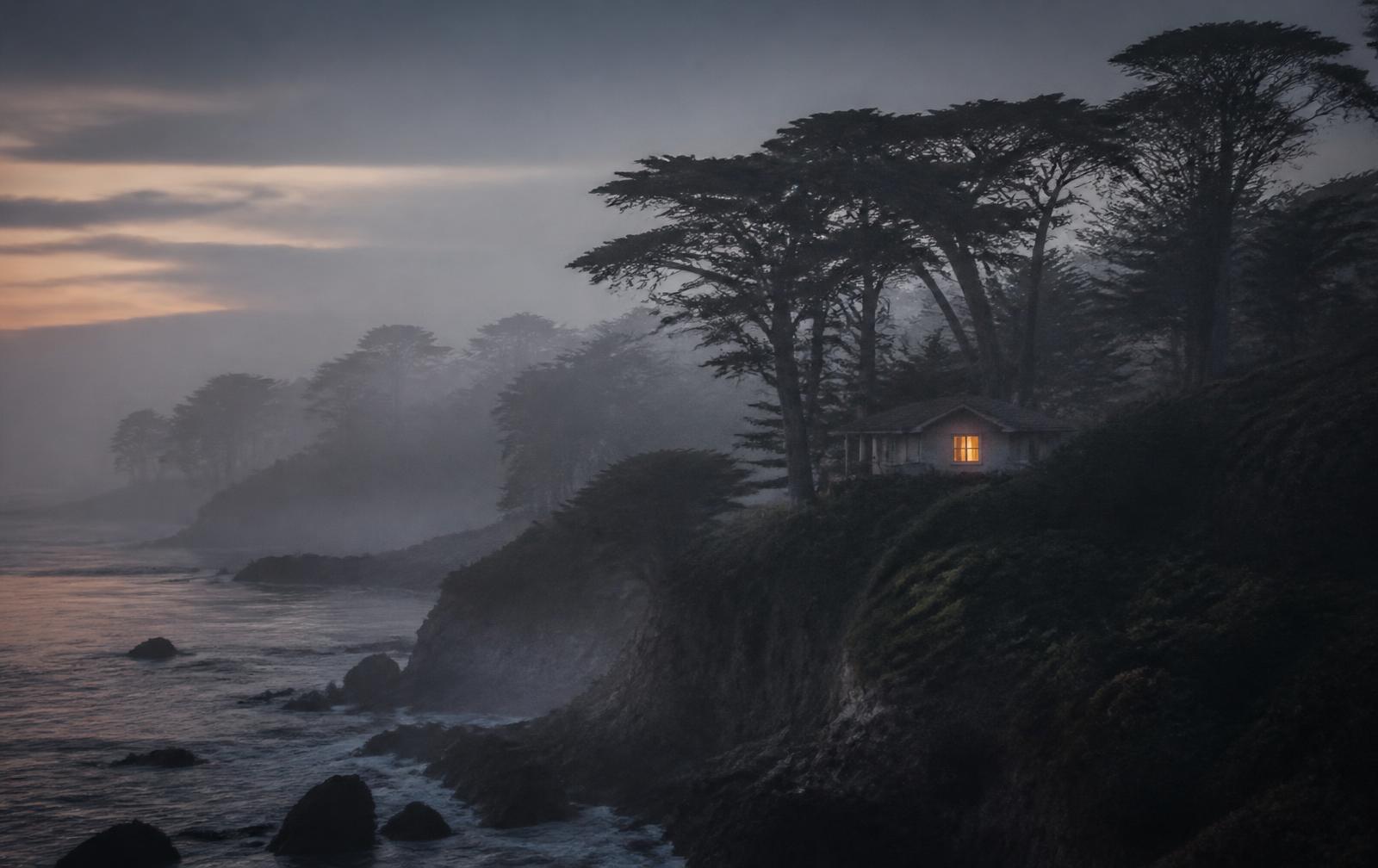 Misty Carmel coastline at dawn with cypress trees and a private cottage glowing softly through the fog