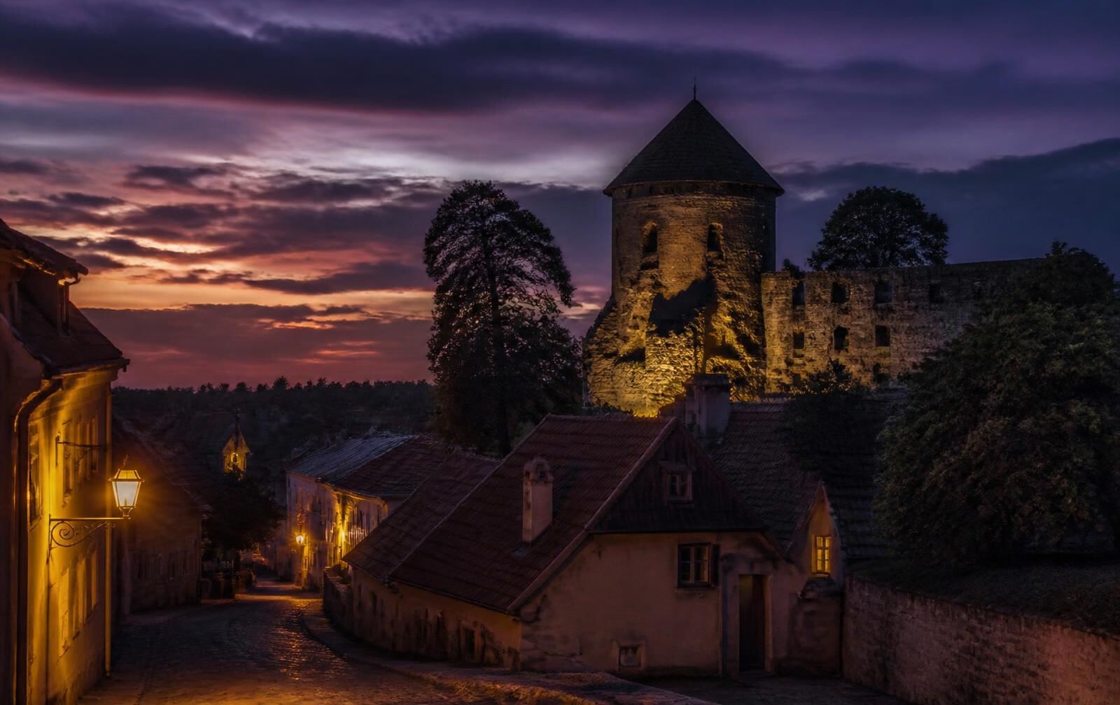 Cēsis at dusk — a medieval stone tower above old rooftops, warm lanterns in a quiet lane under a purple sky