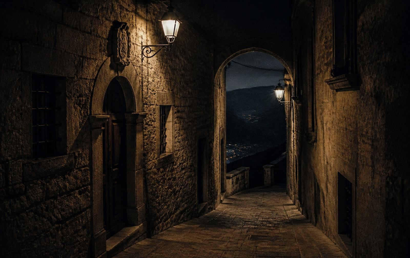 A narrow lantern-lit stone street in the historic city of San Marino at night, opening through an arch onto a dark mountain valley