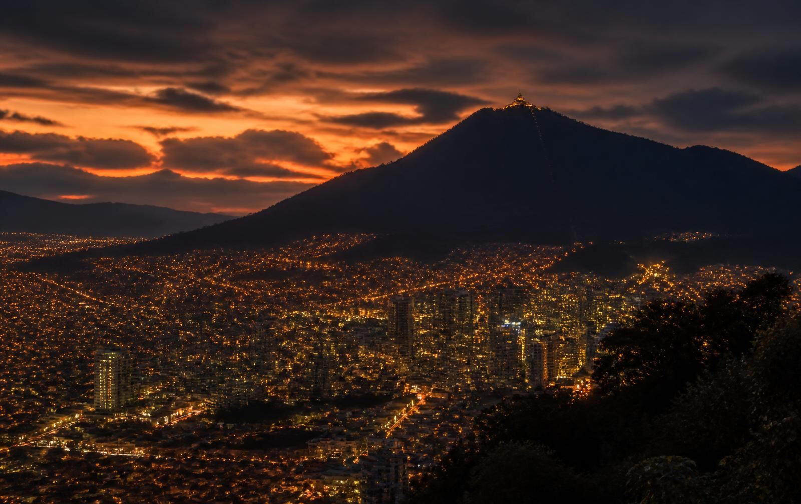 Colombian city lights at dusk beneath a dark mountain silhouette under a dramatic amber sky