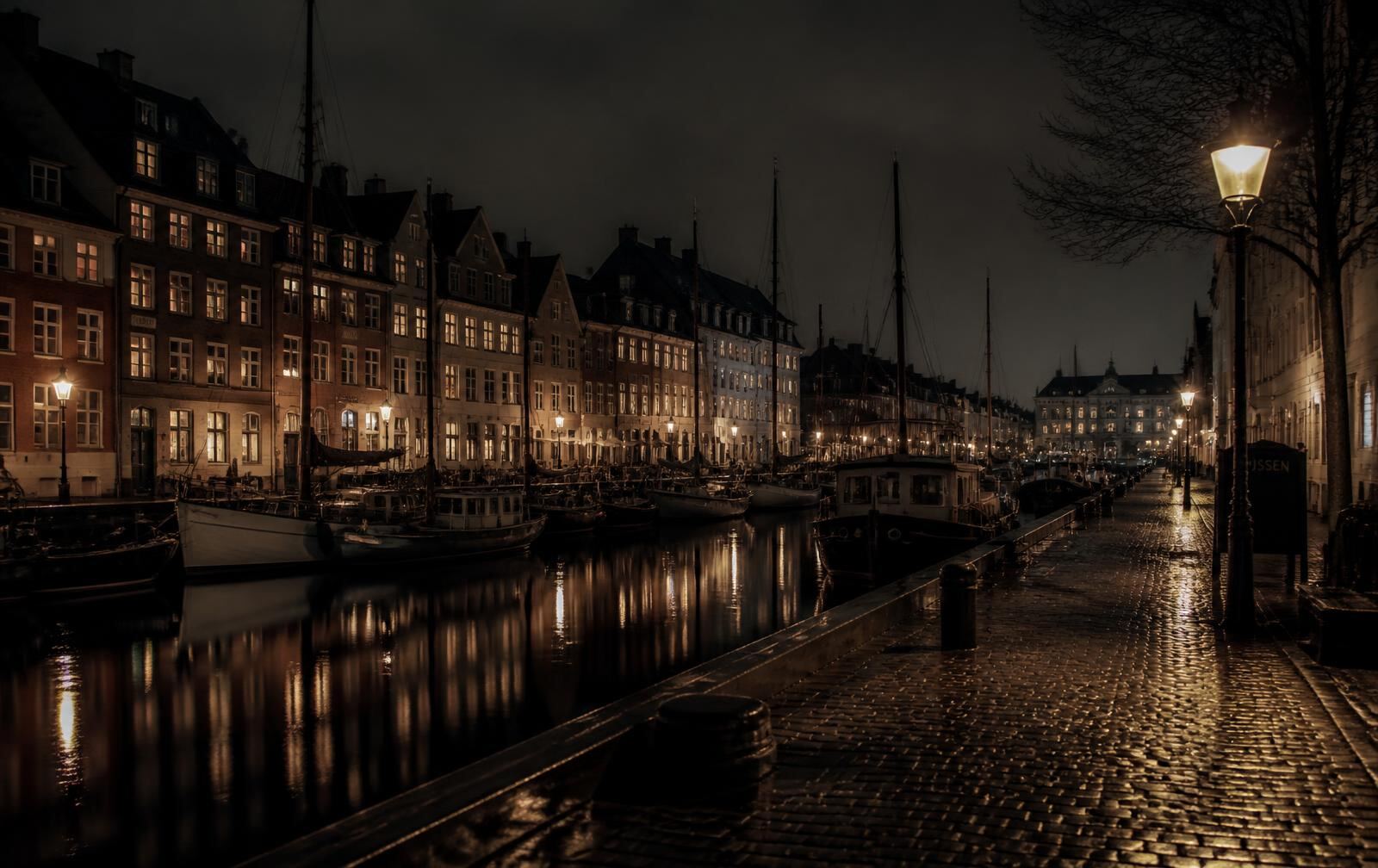 Copenhagen canal at night with old gabled waterfront houses, warm lit windows, moored boats and wet cobblestones reflecting lamplight